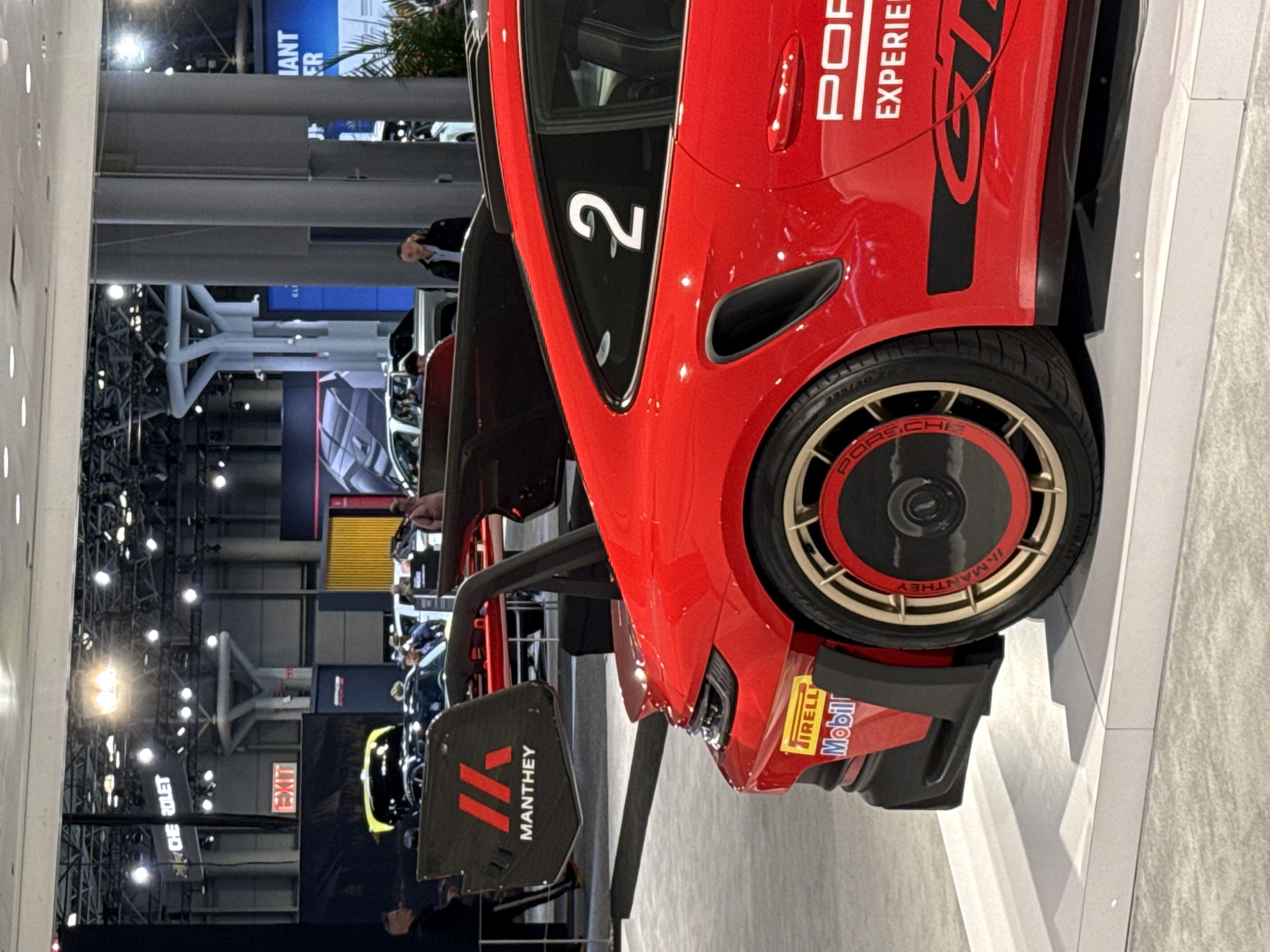 The image shows a red Porsche GT3 race car, adorned with branding from "Porsche Experience" and the number "2". It features distinctive gold wheels with "Porsche" and "Manthey" branding and "Pirelli" and "Mobil" decals on the body. The car is set on a white platform. In the background, there are visible elements of an indoor event, likely a car show, with structures, lighting, and other vehicles partially visible. Overhead lighting fixtures and infrastructure, including beams, suggest the car is in an exposition hall. Elements in the background include signage and structures that imply that this is likely at the New York International Auto Show, which is commonly held in the city of New York, in the United States of America.