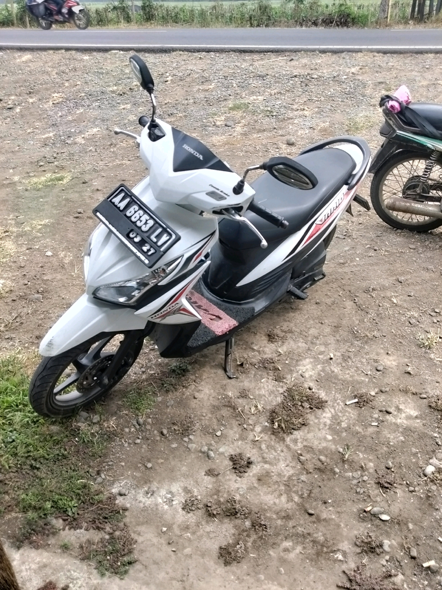 An outdoor, medium-shot, eye-level, full-body photograph shows two parked scooters in the city of Prigelan, Indonesia. The scooter in the foreground is white with black accents and a black seat. It has a license plate that reads "AA 6653 LV 03 27." The scooter in the background is dark green with a black seat and a pink cloth tied to the back. The scooters are parked on a dirt and gravel surface with some patches of green grass. In the background, a paved road can be seen with a blurred image of another scooter in motion, and a grassy field beyond that. The lighting is overcast, and the image appears to have been taken in the daytime.