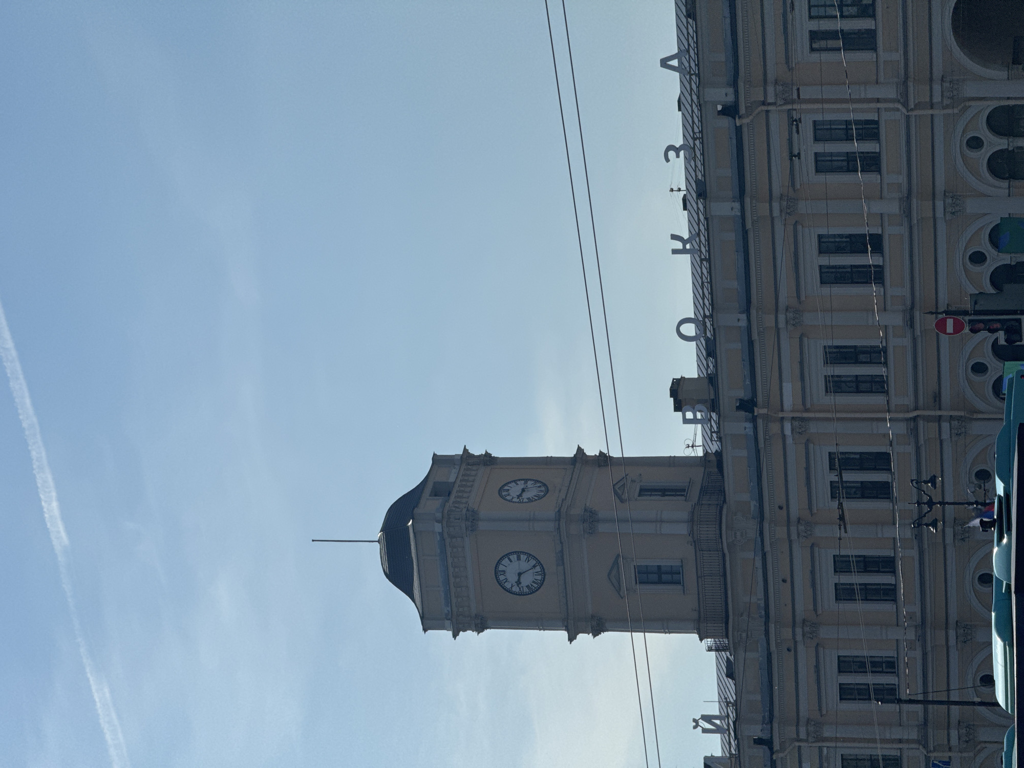 The image depicts the facade of a historic building in Saint Petersburg, Russia, characterized by a prominent clock tower and an inscription above the windows. The building exhibits classical architectural elements, including columns, arches, and ornate detailing. The clock tower has two faces, each displaying Roman numerals. The inscription, partially visible, appears to read "ВОКЗАЛ," meaning "station" in Russian, indicating the building is likely a train station. The sky is clear blue with a few faint contrails from an aircraft. Power lines crisscross the upper portion of the frame, obscuring parts of the building. In the foreground, the roof of a blue bus and a traffic light with a red signal are visible, suggesting an urban street scene. The lighting indicates it is daytime, likely a sunny day.