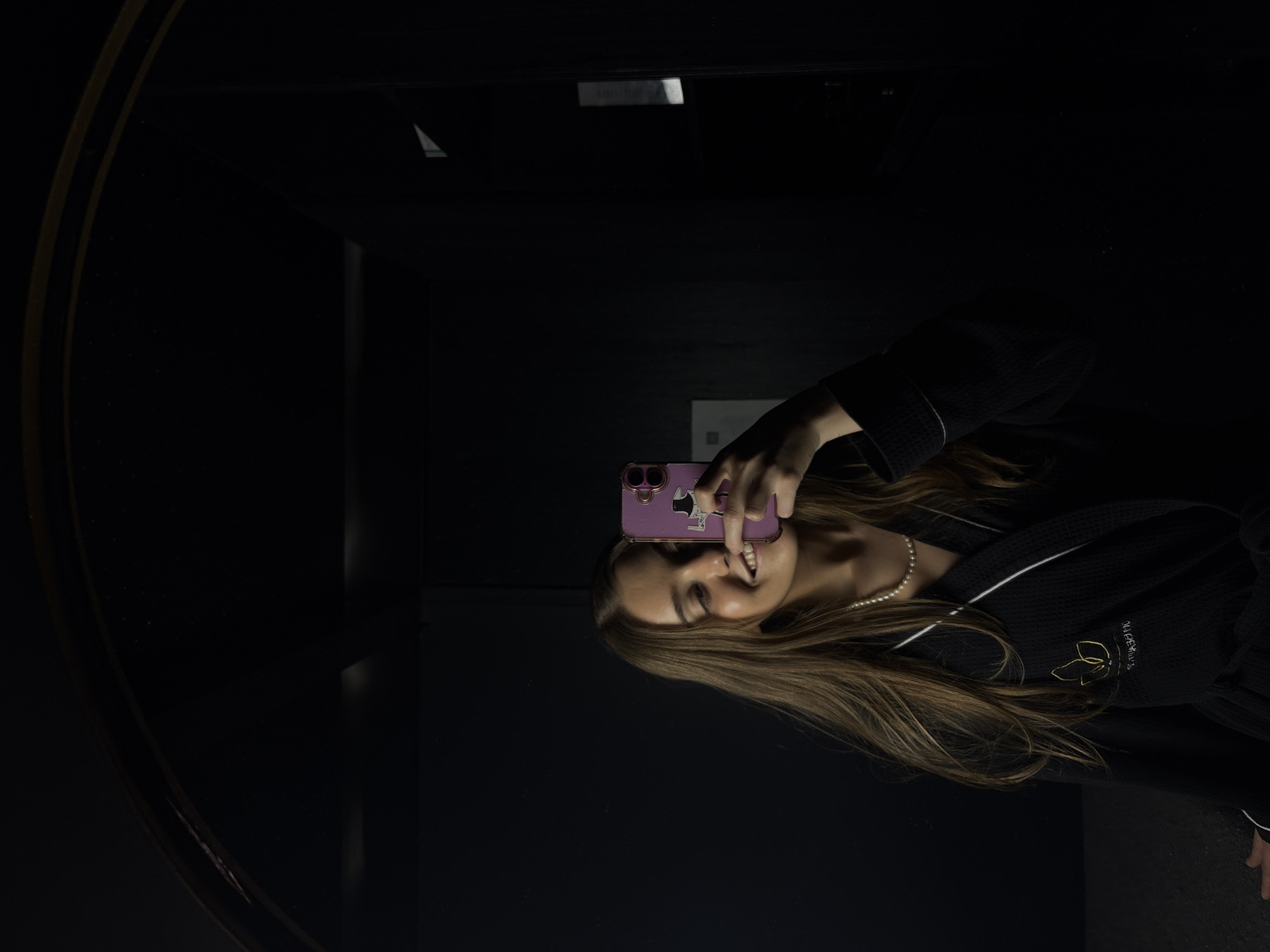 A young woman is taking a mirror selfie in a dark room. She is wearing a black robe with white trim and a pearl necklace. She is holding a pink smartphone with stickers on it in front of her face. The mirror has a dark frame and reflects the dimly lit room, partially revealing architectural details. The location is suspected to be in Ciudad López Mateos, Mexico, but there are no visible clues within the image to confirm the precise location.