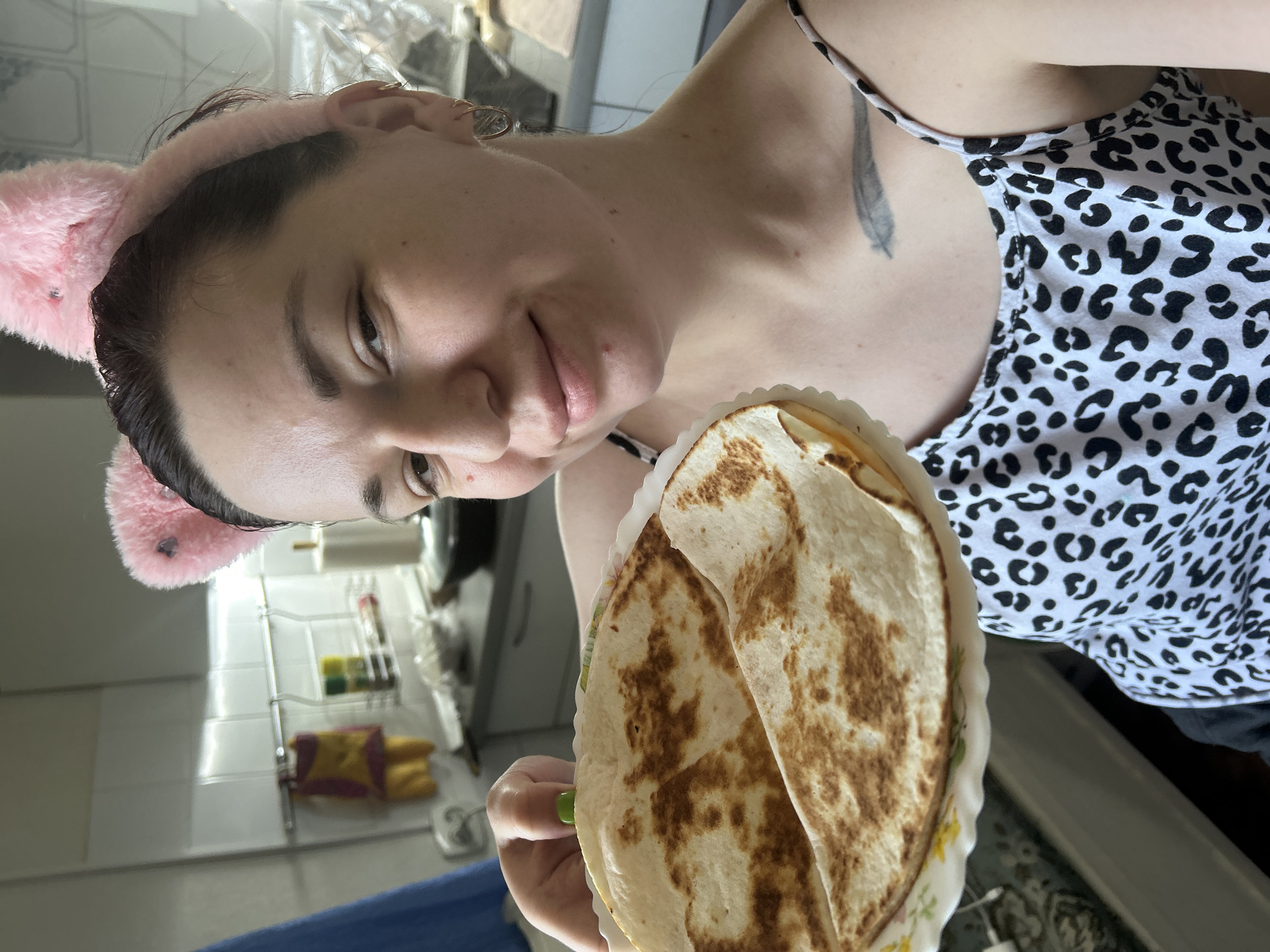 A woman with pink cat ears on a headband smiles as she holds a plate of quesadillas in a kitchen. She is wearing a black and white leopard print tank top. The kitchen has white tiled walls and a rack with kitchen items. The woman has a tattoo of a feather on her chest. The scene is set in an indoor residential kitchen.