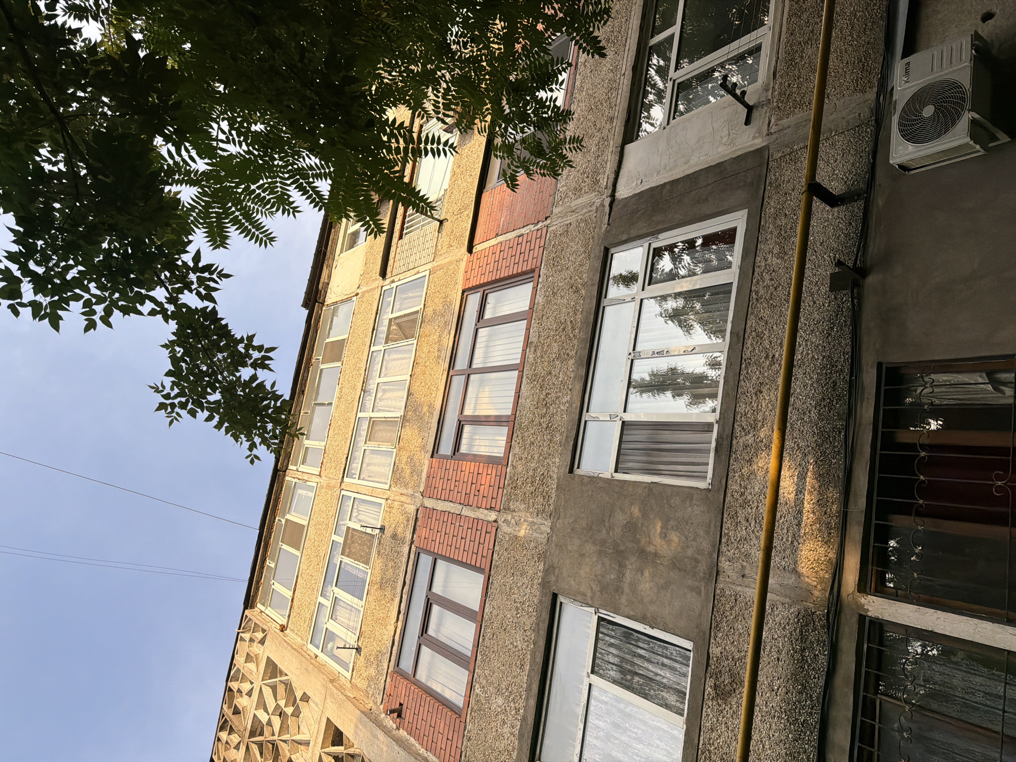 This is a low-angle, upward shot of an apartment building in Shymkent, Kazakhstan, under a clear blue sky. The building's facade is a mix of textured concrete and lighter-colored sections, punctuated by numerous windows. Some sections of the building are decorated with geometric patterns. Lush green tree branches hang from the upper left of the frame, casting dappled shadows on the building.
The windows vary in style: some are large with white frames and blinds, while others have darker frames and appear to have tinted glass. There are also some smaller windows. Visible on the right side of the building is a white air conditioning unit with the brand name "Klima" visible on it. A yellow pipe runs vertically along the building, supported by metal brackets. Electrical wires are also present, stretching across the sky.
The lighting suggests it is either early morning or late afternoon, with the sun casting a warm glow on the building. The overall atmosphere is one of everyday urban life in a residential area. No people are visible in the frame.