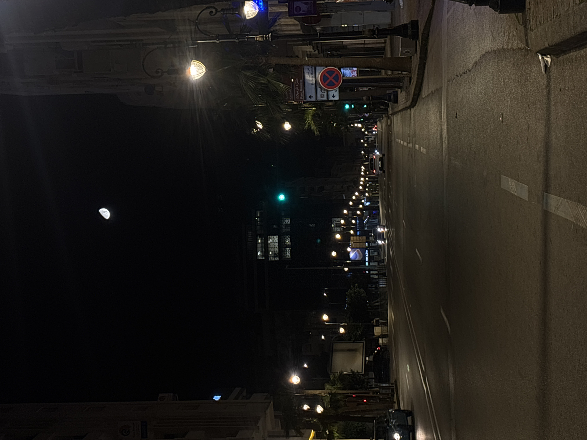 The image depicts a wide urban avenue in Tanger, Morocco, at night. A dark sky dominates the upper portion, featuring a distinct crescent moon in the upper center. The street is illuminated by numerous ornate streetlights emitting warm yellow-orange light, lining both sides of the roadway.
The asphalt road consists of multiple lanes, marked by white solid and dashed lines, and appears to be dry. Traffic is sparse, with a dark-colored vehicle (possibly teal) partially visible in the left foreground, facing away from the viewer with its headlights on. Further down the road, several vehicles with illuminated headlights are approaching from the distant perspective, while others show red taillights moving away.
Along both sidewalks, palm trees are regularly spaced. On the right sidewalk, a multi-directional street sign points towards "Parc Amitié" (straight ahead) and "Palais Marchan" (left). Below this, a circular red sign with a blue background and a red 'X' indicates "No Stopping or Parking." A green traffic light is visible on the right side, mid-distance, with another green light further down the avenue. Buildings of varying heights and architectural styles line both sides of the street, some displaying illuminated windows or commercial signage. Two faint human figures are discernible on the right sidewalk in the mid-ground. A small amount of white debris is visible on the curb in the far right foreground.