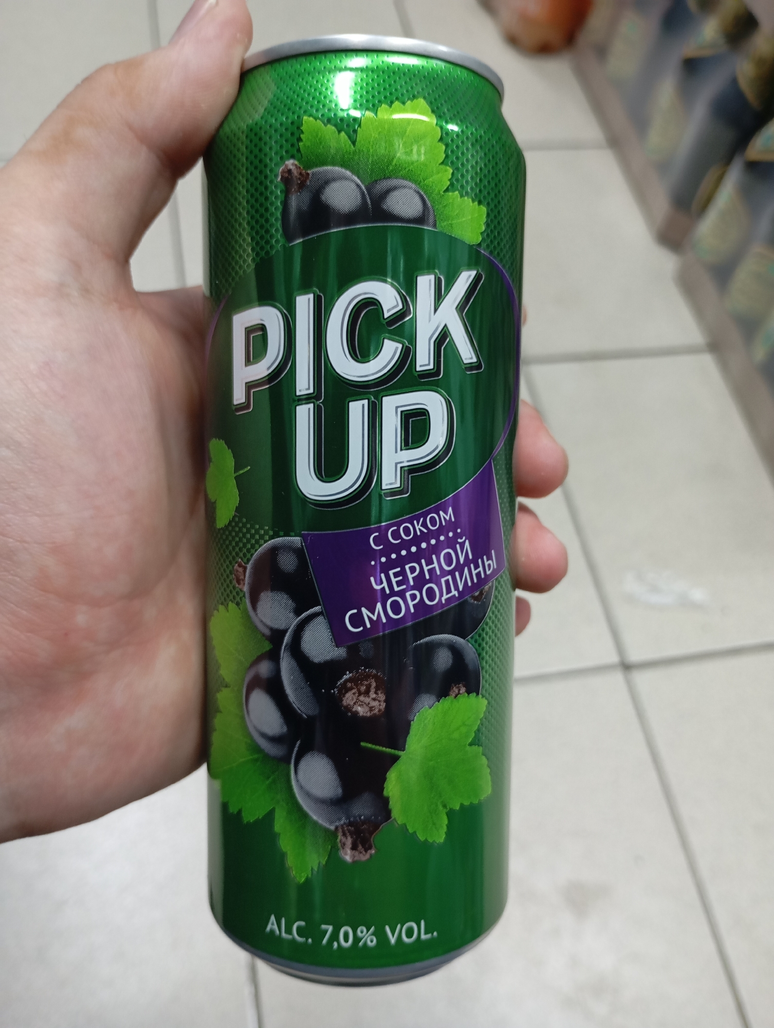 The image shows a hand holding a green aluminum can of "PICK UP" black currant flavored drink. The can has a textured green background with images of black currants and green leaves. The text on the can includes "PICK UP", "С СОКОМ ЧЕРНОЙ СМОРОДИНЫ" (with black currant juice), and "ALC. 7,0% VOL." The background of the image is blurry, showing what appears to be a retail environment with shelves of products and a tiled floor. The scene does not provide any specific information about the location in Moscow, Russia, or any actions or interactions beyond holding the can.