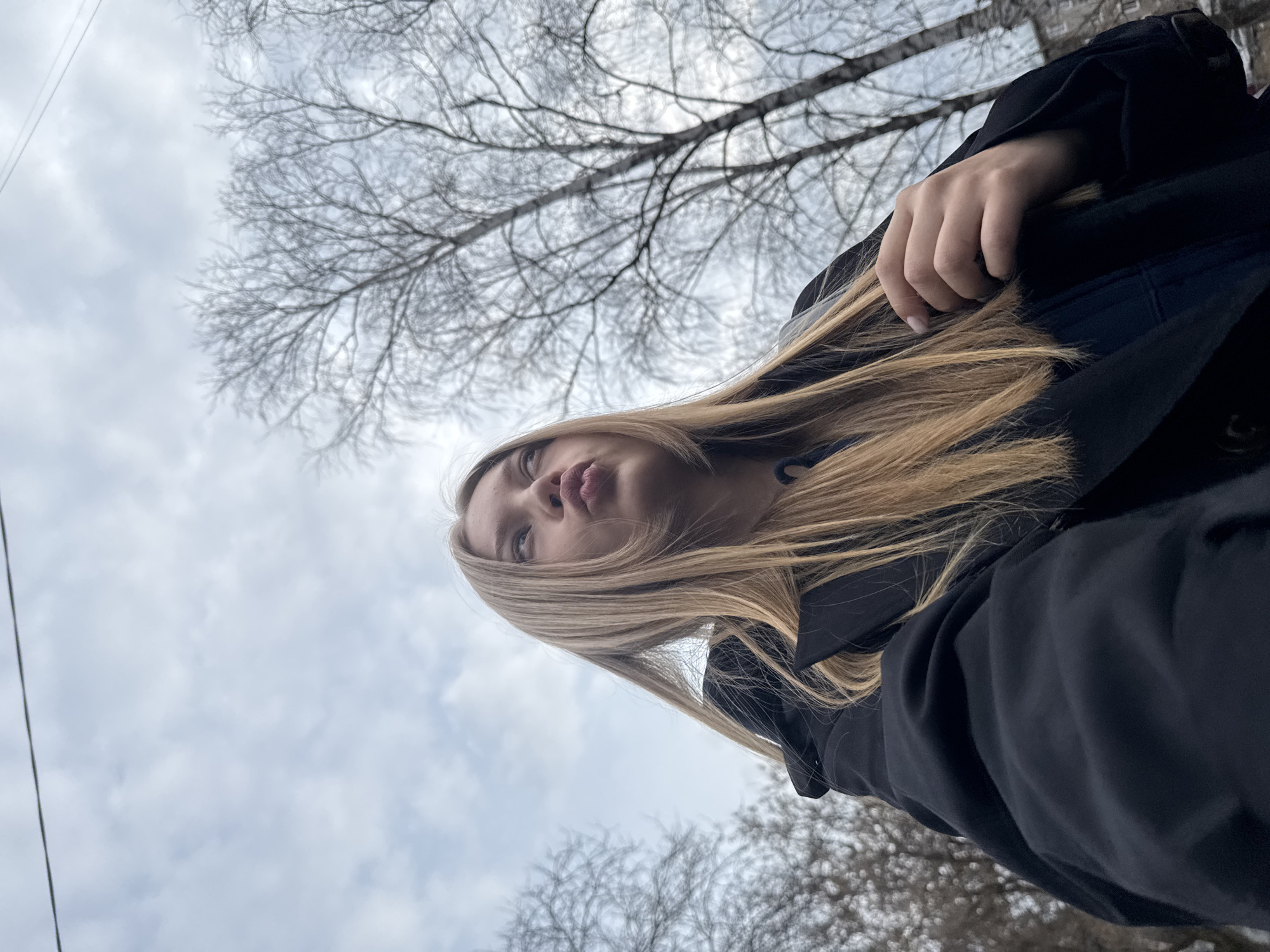 A low-angle shot depicts a fair-skinned young woman with long, light-brown hair. She is wearing a black coat and is posed with pursed lips. Bare tree branches are visible against an overcast, cloudy sky. A building is visible in the background behind the trees. The image appears to have been taken in Chaikovsky, Russia.