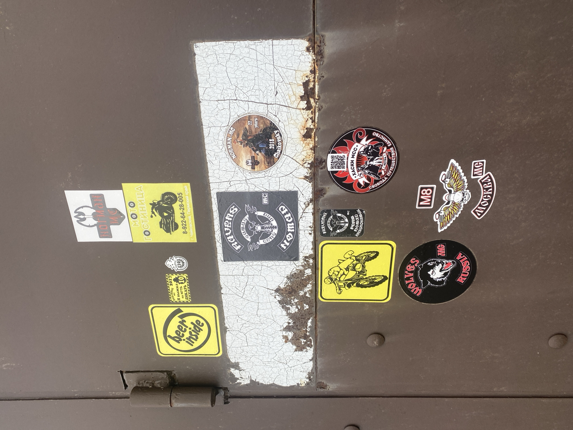 The image displays a weathered, dark brown metal surface, possibly a door or gate, adorned with numerous motorcycle club and event stickers. A prominent vertical seam runs down the center, revealing severely cracked and peeling white paint underneath, alongside significant rust. A metal hinge is visible at the bottom left.
No activity is taking place in the scene; it is a static shot of the decorated surface.
The stickers provide strong location cues, indicating the setting is in Russia, specifically referencing places like "Chaikovsky" and "Permskiy kray" (Perm Krai), "Krasnokamsk," "Istra," and "Moscow." Many stickers feature motorcycle motifs, club names, and event details.
Visible text includes:
- Yellow stickers: "beer inside," "RAVERS 2017 Я выжил ДИКАРЬ 10" (I survived the wild/savage 10), "MOTO ГОСТИНИЦА" (MOTO HOTEL) with a phone number "8-922-64-28-005" and address "г. Чайковский, Пермский край," and one depicting a dirt bike.
- Circular sticker: "NORMAN-MC 2018 Chaikovsky" with a motorcycle rider, and another small one stating "22 years."
- Black square sticker: "Nomad Riders MC Ravens Krasnokamsk" featuring skulls and a motorcycle.
- Circular black sticker: "ХАСКИ МСС. • ИСТРА СВОБОДА ПУТЕШЕСТВИЯ ЕДИНСТВО" (HUSKY MCC. • ISTRA FREEDOM OF TRAVEL UNITY) with a QR code.
- White and black sticker: "M8 Москва MG" (M8 Moscow MG) with a winged engine design.
- Circular black sticker: "WOLVES MC RUSSIA" with a wolf head.
- Other smaller stickers referencing "Ravens MC Perm" and "PERM NOMAD."
The collection of stickers reflects a vibrant motorcycle culture, marking affiliations and participation in various events across different Russian locations.