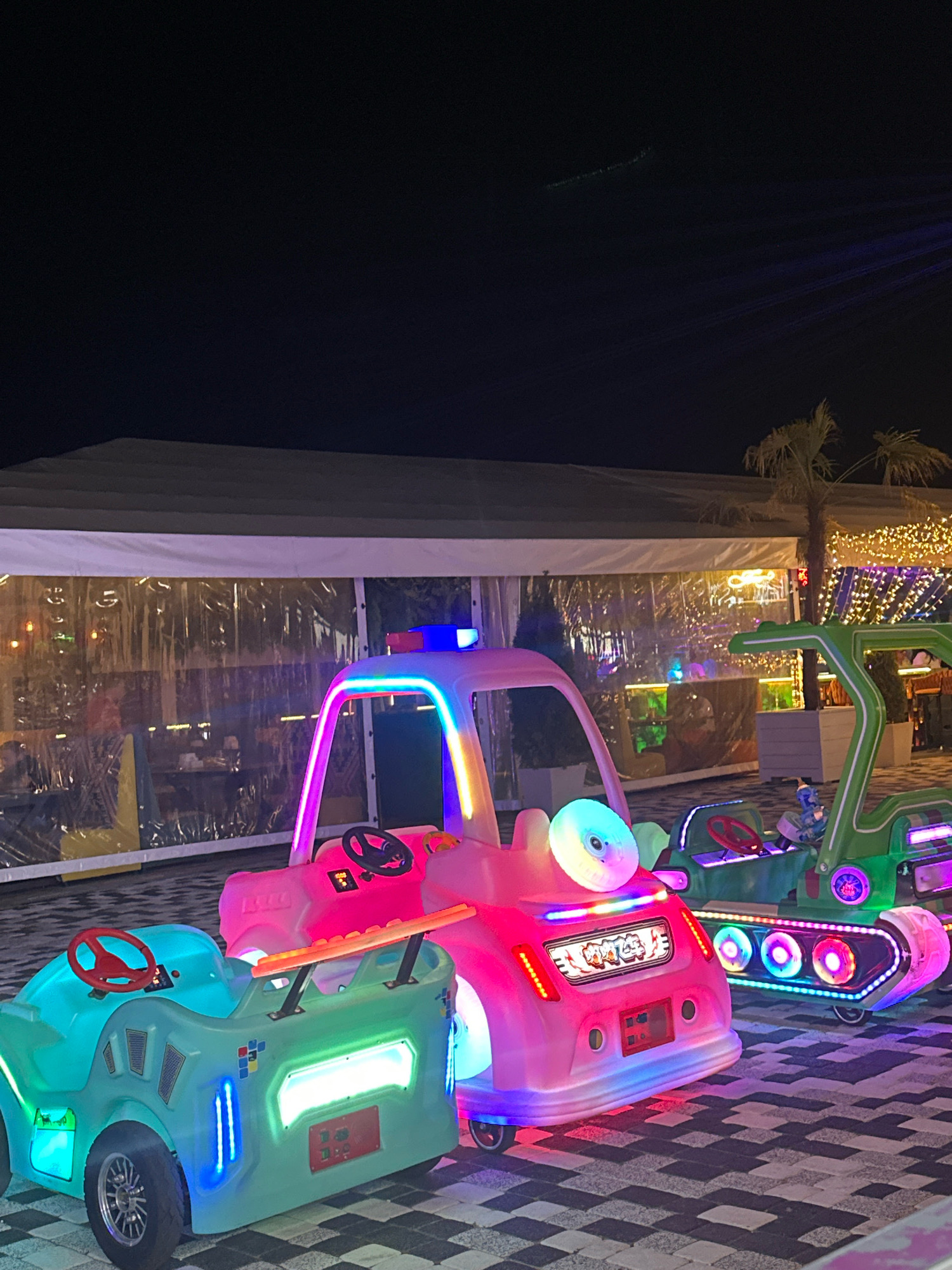 This is a night-time outdoor photograph of three illuminated children's amusement ride vehicles parked on a patterned tiled surface. The vehicles are a light blue car, a pink car, and a green military-style vehicle. The pink car has a police car motif with a blue flashing light on top. The vehicles are brightly lit with colorful LED lights. Behind the vehicles is a large, white tented structure with clear plastic walls, revealing tables and chairs and people seated inside. String lights are visible hanging from the ceiling of the tent and further back, a palm tree and more decorative lights are present. The background sky is dark. The scene appears to be located in the city of Lermontovo, Russia, likely at an amusement park or entertainment venue.