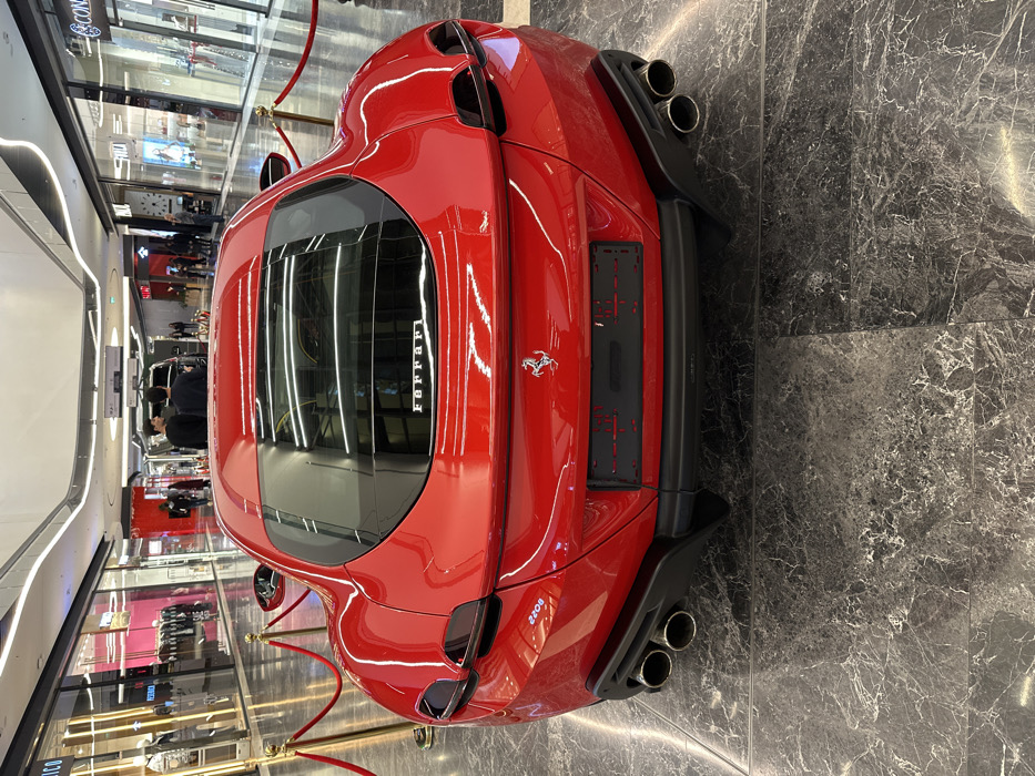The scene captures a red Ferrari sports car, specifically a Ferrari Roma, displayed prominently from the rear within a modern, upscale shopping mall. The car features a glossy red finish, quad circular exhaust pipes integrated into a black rear diffuser, and a rear windshield bearing the "Ferrari" script. A silver Ferrari prancing horse emblem is affixed below the rear trunk line, above an empty black license plate holder. The car is cordoned off by a perimeter of gold stanchions linked with red velvet ropes. The floor consists of large, polished dark marble or granite tiles with distinct white veining, reflecting the overhead lighting and portions of the car.
In the background, a brightly lit mall aisle is visible, lined with multiple luxury retail storefronts. To the immediate left, a store window with "VERSACE" reflected text displays mannequins dressed in fashion apparel. To the right, other store facades include "CERRUTI 1881" and "FALDI" signage, showcasing items such as watches and jewelry. The ceiling features modern, curvilinear white panels and integrated linear LED lighting. Several individuals are present in the background, walking or standing. Two men in dark clothing are positioned further down the aisle, near a dark SUV or large vehicle. The environment is illuminated by consistent artificial lighting, producing noticeable reflections on all polished surfaces.
This location is identified as Tashkent City Mall in Tashkent, Uzbekistan.