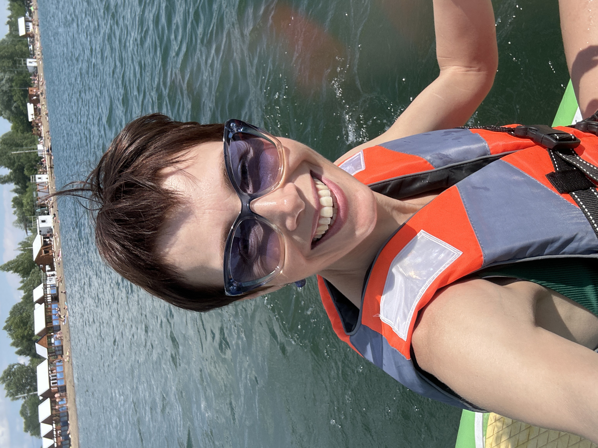 A woman is taking a selfie while on a paddleboard on a lake. She is wearing sunglasses, a life vest, and appears to be smiling. In the background, there are several small beach huts and trees, with a hazy shoreline. The water is calm and choppy. The scene appears to be a sunny day at a lake in Kemerovo, Russia.