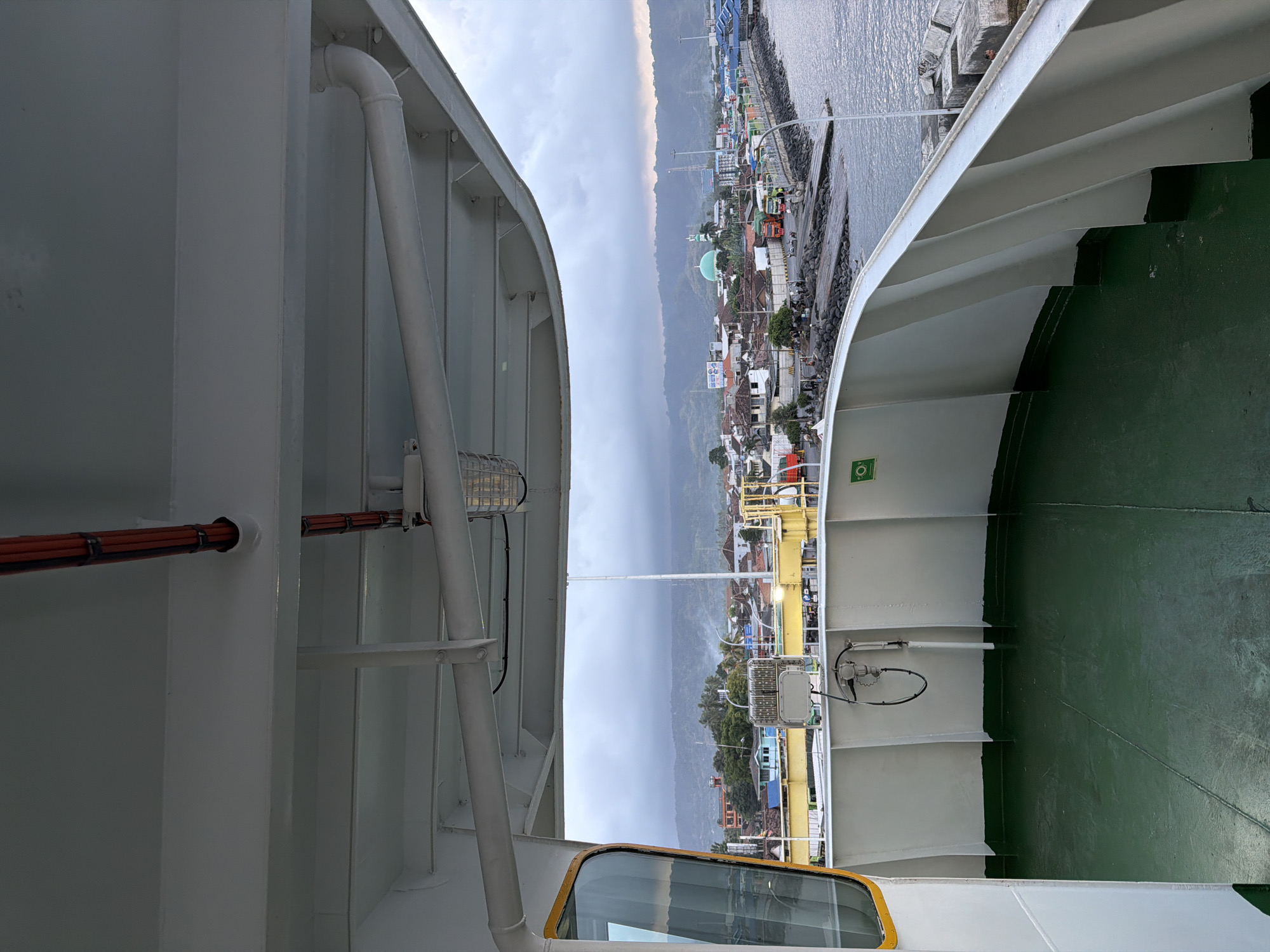The image depicts a view from the deck of a ferry in the city of Ketapang, Indonesia. The foreground shows the green-painted deck of the ferry with white curved railings. Above, an overhang structure supports pipes and lighting fixtures. Beyond the ferry, a harbor area is visible with a concrete pier, several vehicles including trucks, and buildings lining the shore. In the background, a town with various structures, including a prominent green-domed building, is nestled against a backdrop of rolling, cloud-covered hills. The sky is overcast, suggesting a cloudy day. No people are clearly identifiable in the image.