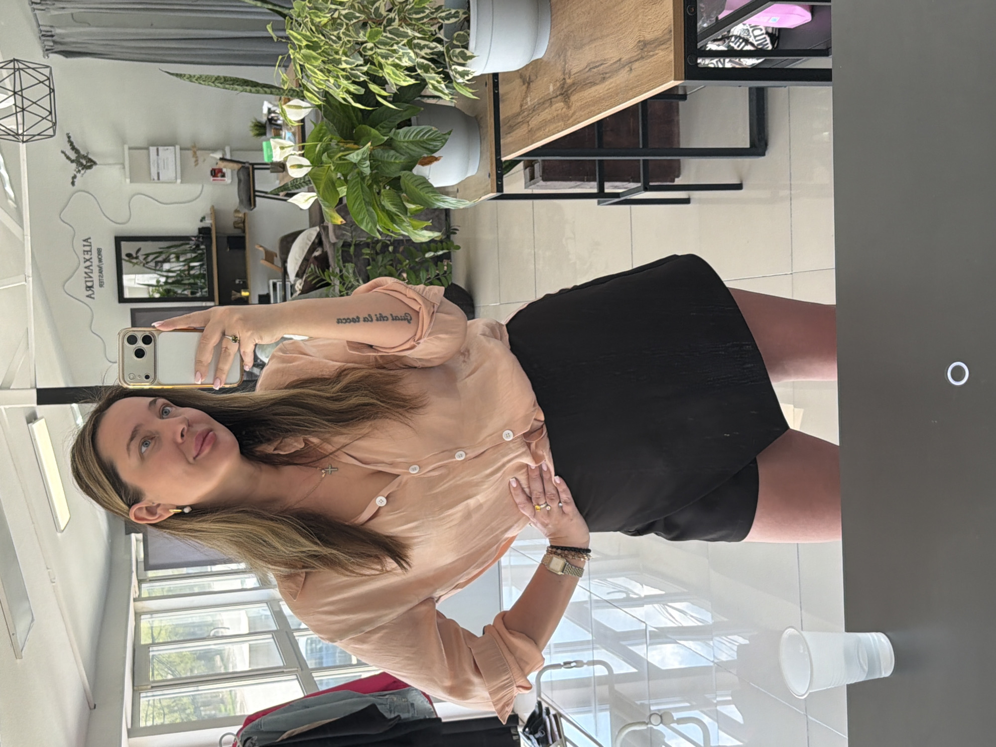 A woman is taking a selfie in a room that appears to be a beauty salon or aesthetician's office. The room has light-colored tile flooring, large windows, and a gray wall. A table with plants on top is positioned in the background. The woman is wearing a peach-colored button-down shirt, a black skirt, and jewelry. A disposable cup is attached to the wall. There is a sign on the wall with the name "ALEXANDRA". Although the city of Chaikovsky, Russia is specified, there is nothing definitive in the image to support or contradict this claim. The image mainly depicts the woman and the interior of the room.