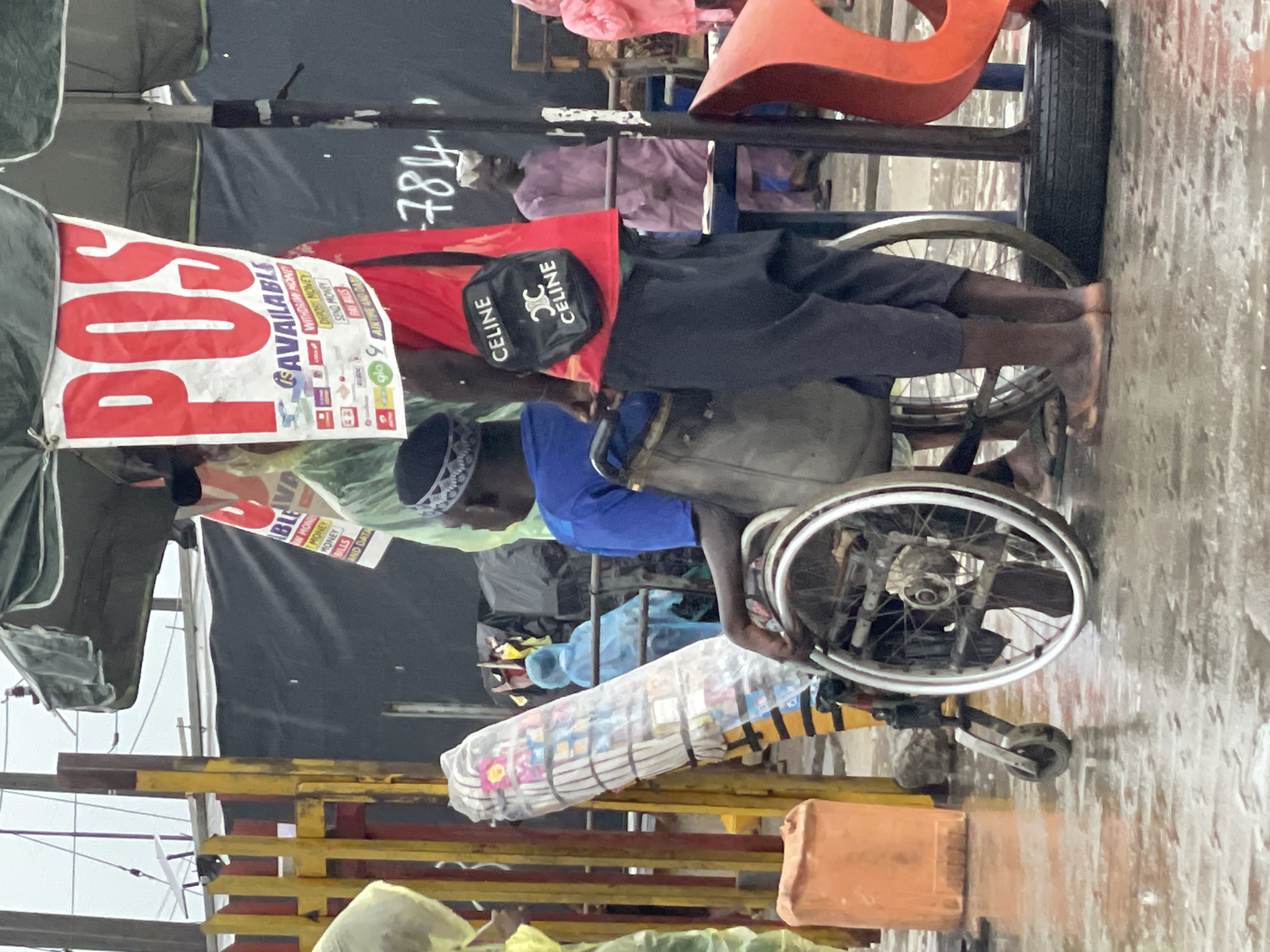 An individual in a wheelchair, identified by a small black cap and blue shirt, is seen in a bustling outdoor area, likely a market, in Lagos, Nigeria. The individual is wearing a black "CELINE" branded bag and is positioned next to a sign indicating POS (Point of Sale) services are available. The sign lists financial transactions such as withdrawing, depositing, sending money, and paying bills, implying the individual may be offering these services. The wheelchair is loaded with wrapped goods. The environment includes various market stalls, fabric covers, and general clutter, indicative of an informal commercial setting.