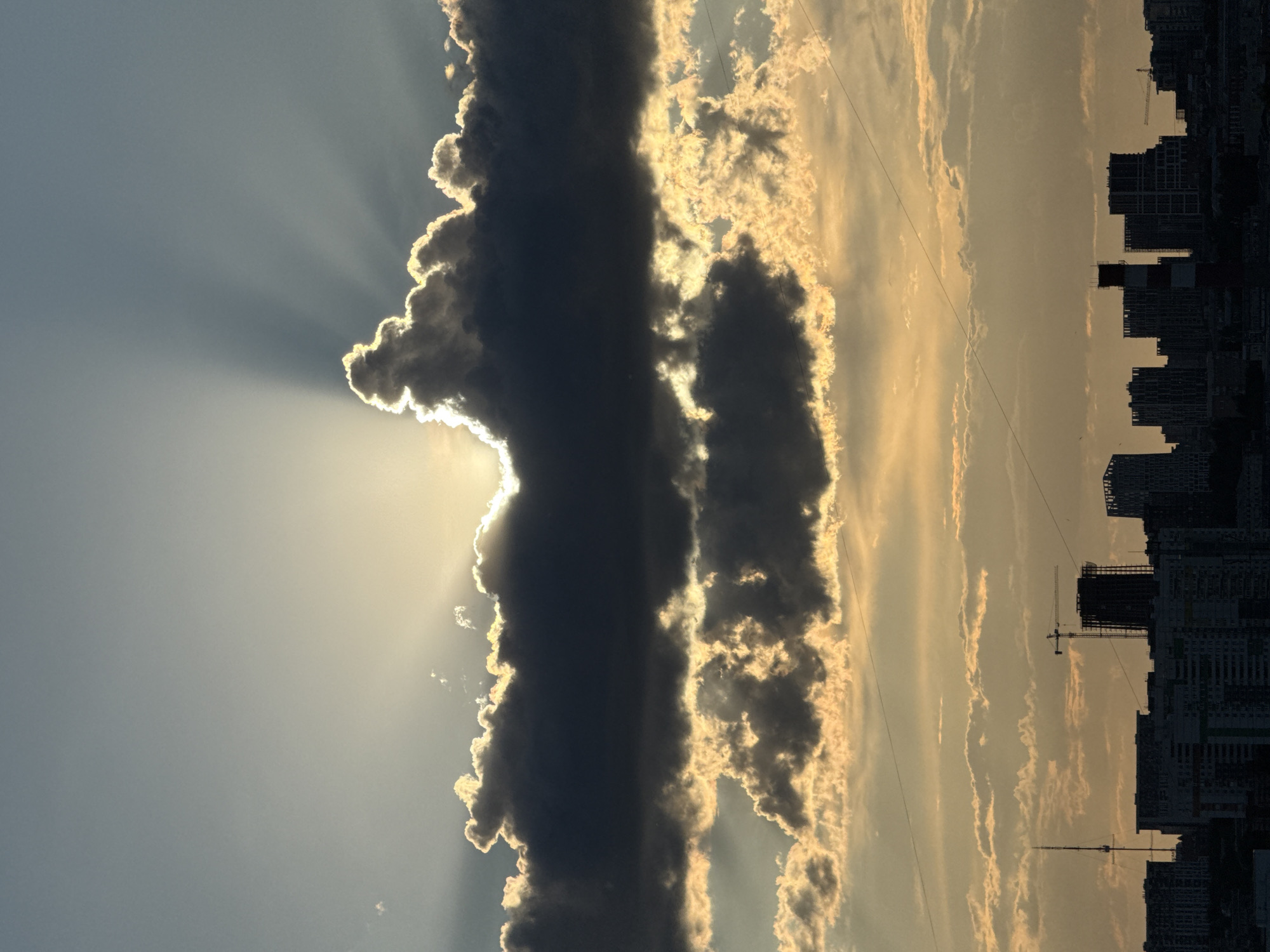 The image depicts a dramatic sky over a cityscape. Dominating the left side is a large, dark cloud formation with a bright rim, backlit by the sun, creating sunbeams radiating outwards. To the right of this cloud, thinner bands of clouds stretch horizontally across the frame. The silhouette of buildings is visible on the right edge of the image, suggesting an urban setting. Given the request for a location, it is a view overlooking a section of Izhevsk, Russia. Specific details about people or actions are absent in the image. Objects include clouds, buildings, and potentially a construction crane. The environment suggests either sunrise or sunset, with a strong contrast between light and shadow.