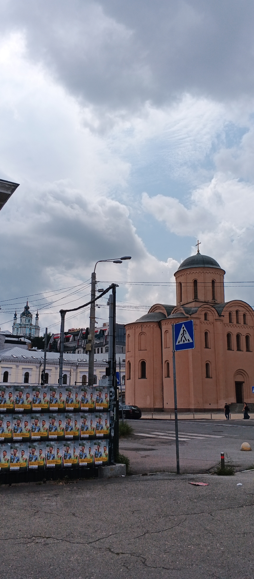 The image depicts a street scene in Kyiv, Ukraine, focusing on a large, peach-colored domed building that appears to be a church or religious institution. In the foreground, a series of posters are displayed on a wall, featuring portraits of multiple men. To the right of the posters, a pedestrian crossing is marked on the asphalt, with a blue and white sign indicating the pedestrian crossing. A black car is parked near the building, and several people are visible in the background, walking near the entrance of the building and across the street. Further in the distance, another distinct blue-domed church can be seen. The sky is overcast with heavy clouds, suggesting a possibly rainy or stormy day. Electrical wires and a lamppost are prominent elements in the middle ground, indicating an urban setting. The overall impression is that of a public space in a historical area of Kyiv, with elements of both religious architecture and everyday urban life.