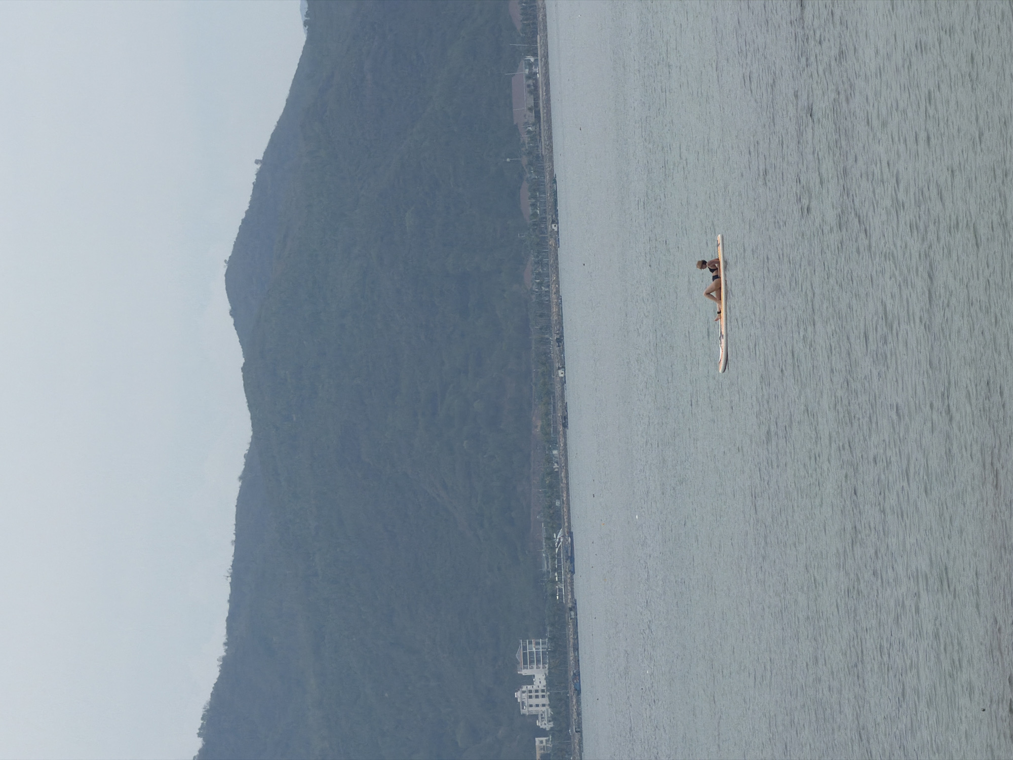 The image shows a landscape of Bac Nha Trang Ward, Vietnam, featuring a large mountain on the left and a body of water occupying the right side of the frame. Along the shore between the mountain and water, there is a strip of buildings and greenery, indicating a developed area. In the middle of the water, a person is seen on a paddleboard.