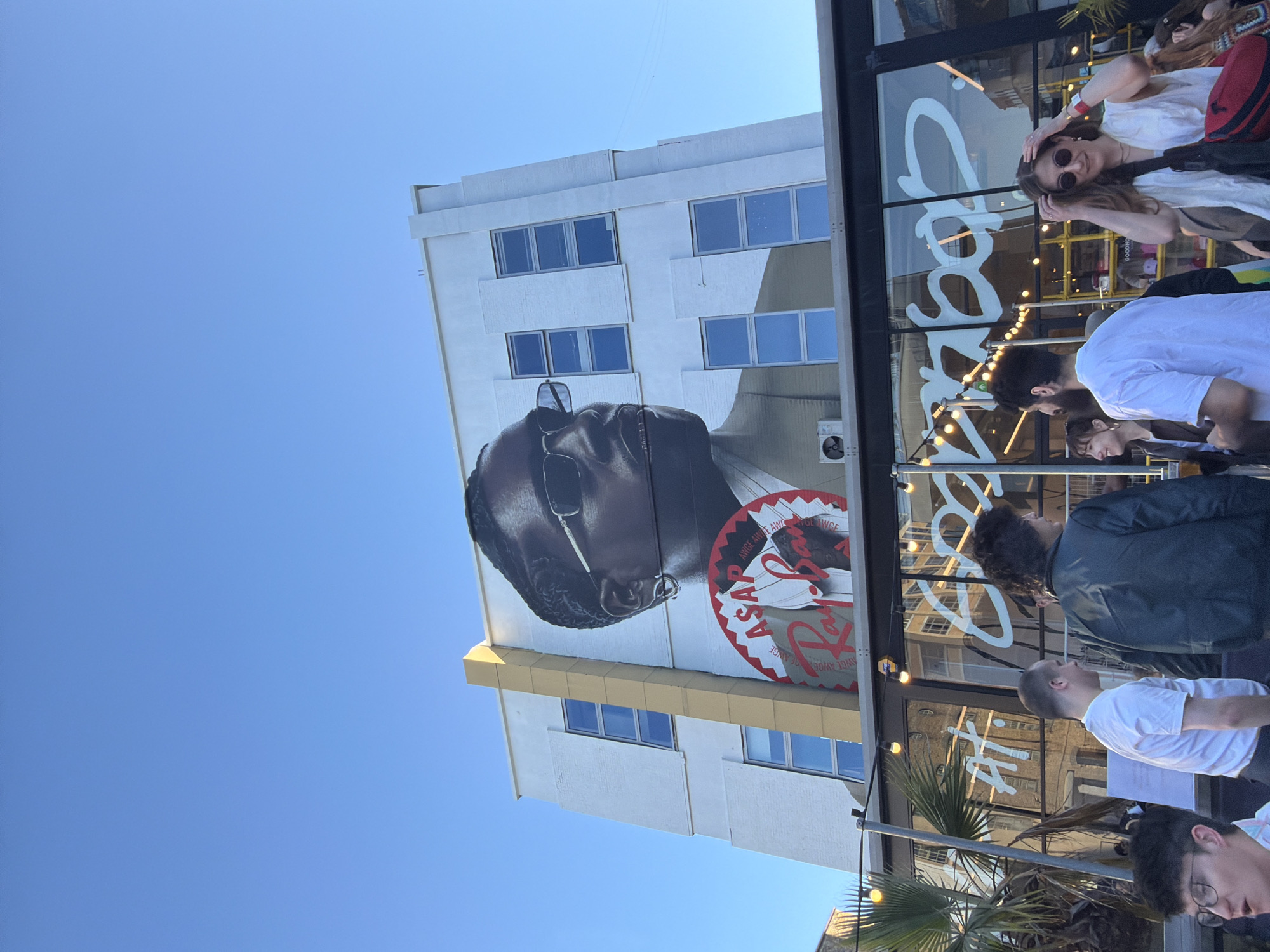 The image captures an outdoor scene in London, United Kingdom. A multi-story white building features a large mural of A$AP Rocky wearing sunglasses and a Ray-Ban logo. Below the mural are a set of windows and what appears to be an air conditioner. Adjacent to the building is a storefront with large windows displaying "Roots." People stand outside the storefront, including a woman with sunglasses, wearing a white top, a man with dark curly hair, and other people in casual attire. In the foreground are palm trees. The sky is clear and blue.