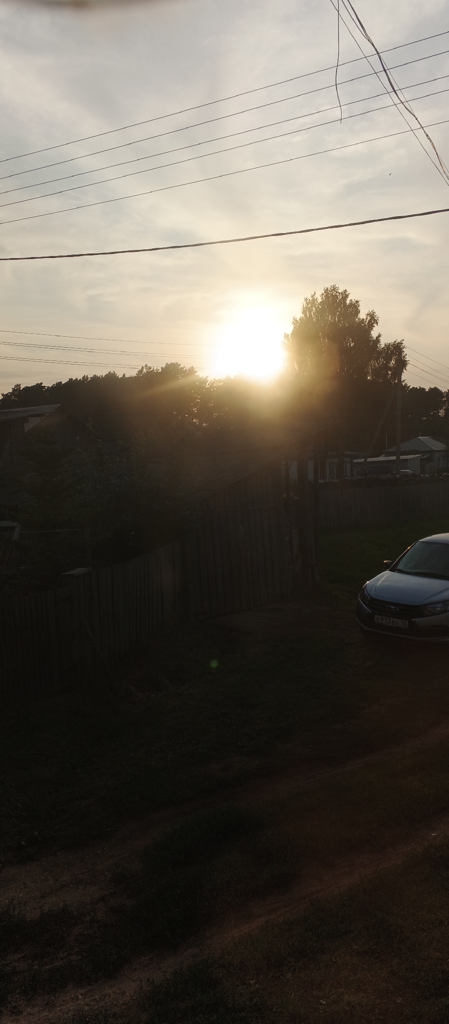 The image shows a rural scene at sunset. The sun is low on the horizon, casting a bright glare and silhouetting trees and buildings. In the foreground, there is a wooden fence and a grassy area. A silver car is parked on the right side of the image. Overhead, power lines are visible against the sky. The scene appears to be in the village of Podgornoye, Russia.