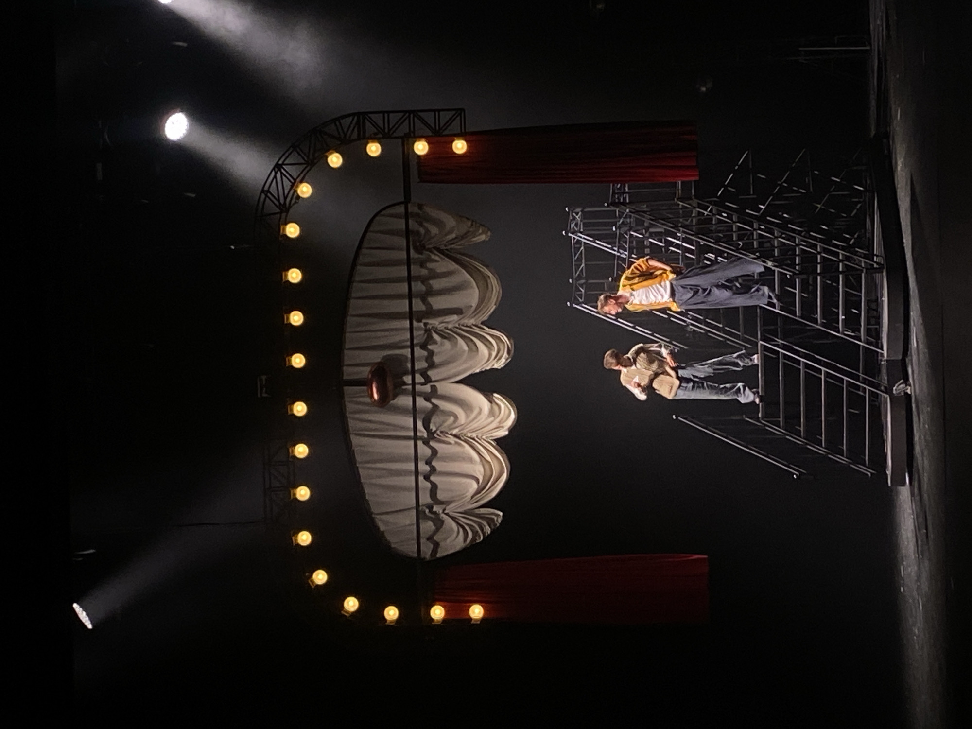 Two people are standing on metal scaffolding on a stage. One person is wearing a yellow jacket, and the other is wearing a brown jacket. In the background, there is a large semi-circular structure with lights around the edge. There are also two red curtains. The location is Заволжье, Russia.