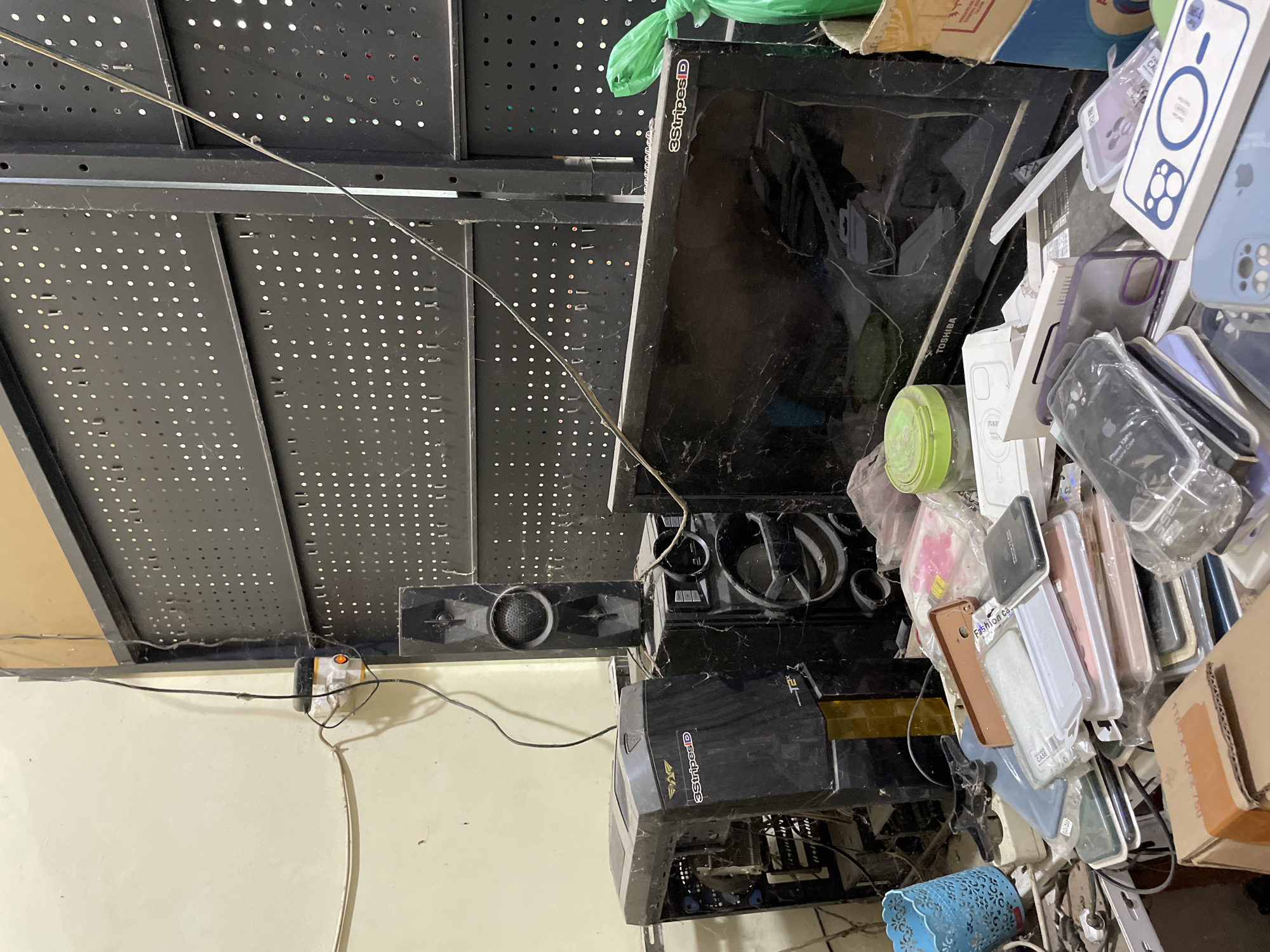 The image depicts a cluttered desk or workstation in what appears to be a workshop or storage area in Pasar Paloh, Indonesia. The scene is dominated by electronic equipment and various accessories. In the background, a large pegboard structure is visible, with wires hanging down.
Prominently featured is a black computer tower case, covered in dust and cobwebs, to the left of a two-piece speaker system. The speaker system consists of a taller, thin speaker unit stacked on top of a larger speaker unit. A black monitor, also dusty, is positioned to the right of the speakers, with a green plastic bag partially obscuring its right side. The monitor screen shows signs of damage or cracking.
In the foreground, a significant pile of mobile phone cases is arranged on the surface. These cases vary in color and material, with some still in their original packaging. Other small electronic accessories and miscellaneous items are scattered amongst the phone cases. A blue plastic organizer is visible to the far left. A cardboard box is partially visible at the bottom right corner. The overall impression is one of disarray and neglect, possibly indicating a repair shop, an electronics recycling area, or a storage space for unsold inventory.