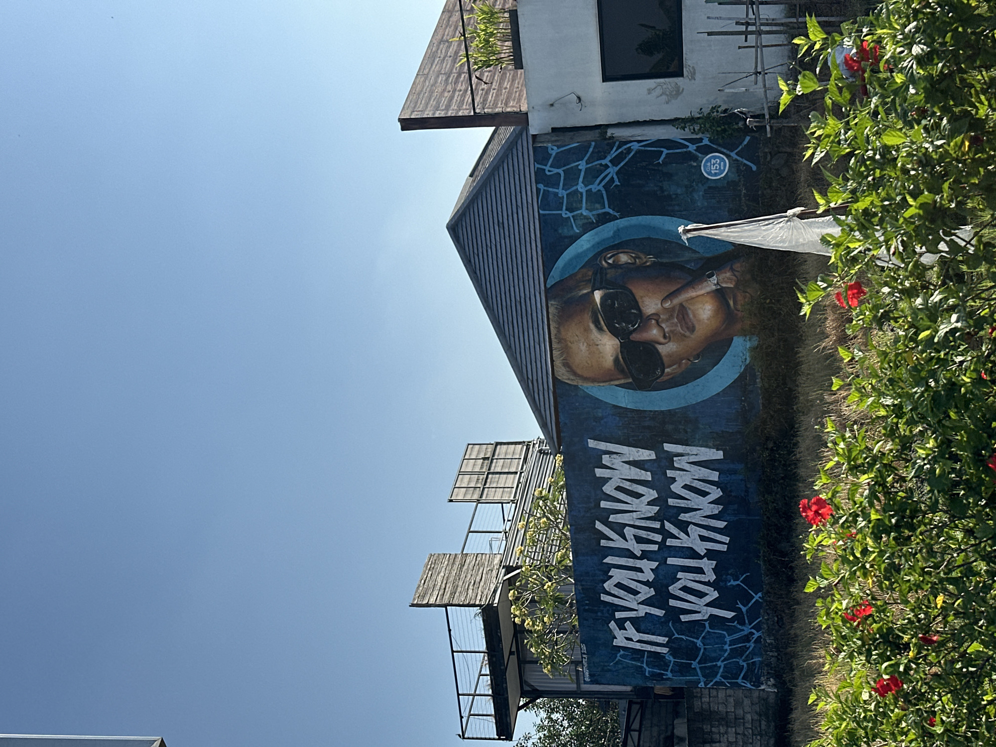 The image captures a mural on the side of a building in North Kuta, Indonesia. The mural features a close-up of a person's face, wearing sunglasses and looking directly at the viewer. Below the face, the text "IF YOU KNOW YOU KNOW" is written in bold, white lettering against a dark blue, abstract background with a crackle pattern.
The building itself appears to be a multi-story structure with some architectural elements suggesting it might be a cafe, restaurant, or boutique hotel. Wooden louvers and balconies are visible, as is a section of white wall with a dark-framed window. The scene is bathed in bright sunlight, indicating it is daytime. Lush green foliage with several red hibiscus flowers is in the foreground, to the right of the building, adding a touch of natural beauty to the urban setting. The sky is a clear, vibrant blue.
The overall impression is one of a vibrant and stylish location, likely a popular spot given the prominent and artistic mural. The phrase "IF YOU KNOW YOU KNOW" suggests an insider reference or a sense of community among those familiar with the place or its significance. The weather appears to be sunny and pleasant, typical for Indonesia.