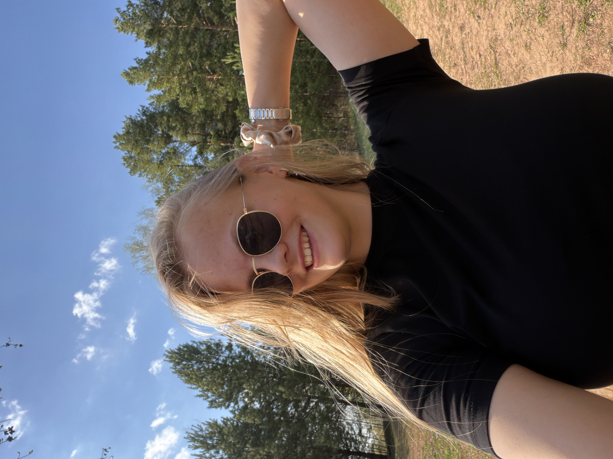 A fair-skinned person with long blonde hair is smiling and wearing sunglasses and a black top. The person is outdoors, likely in a park or wooded area. The sky is blue with some clouds. Trees are visible in the background. The exact location within the city of Kamennoye, Russia, cannot be determined from this image.