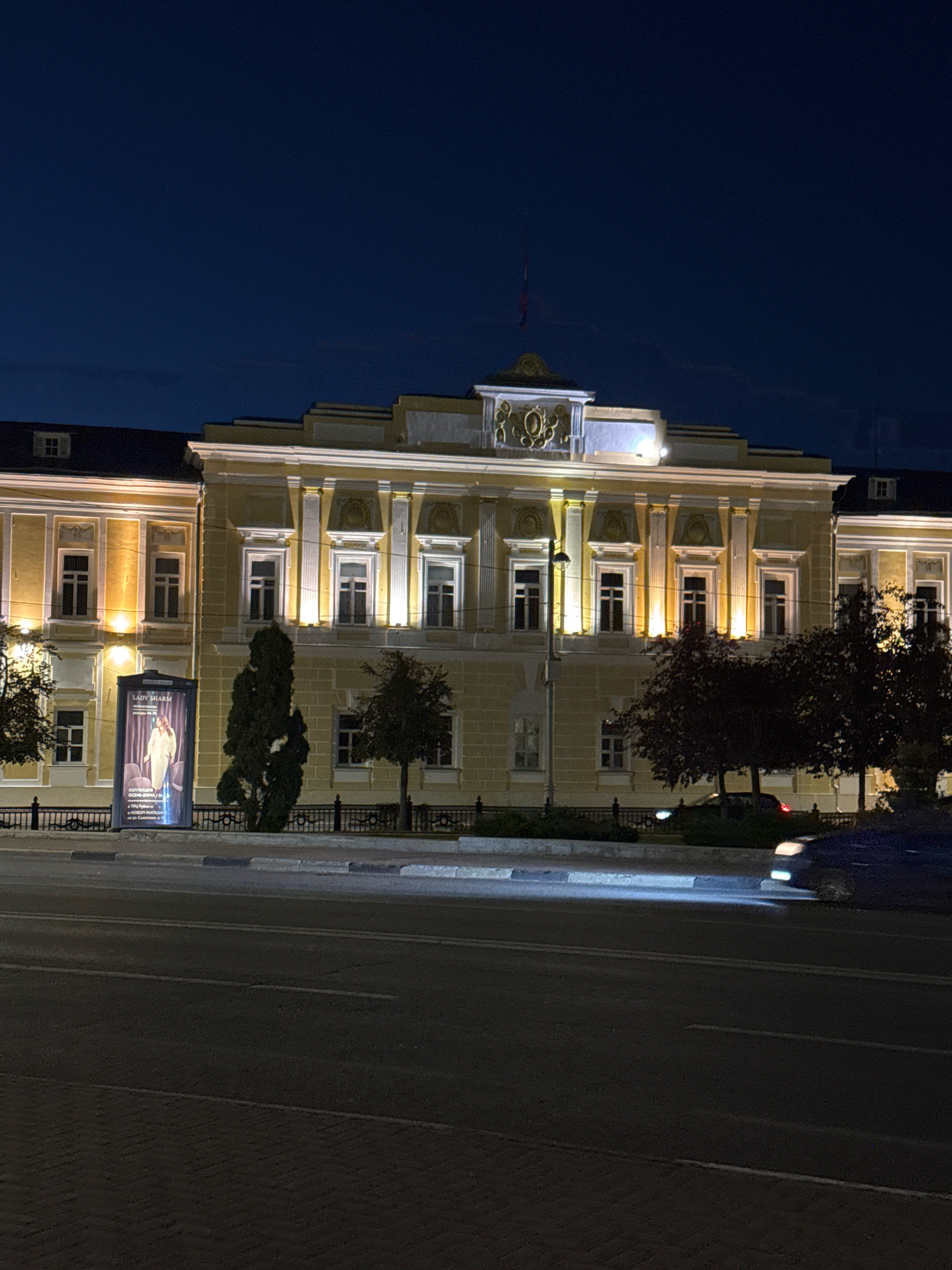 The image shows a building illuminated at night in Tver, Russia, likely on a street with traffic. The building, a historical structure painted yellow with white architectural details and a dark roof, is symmetrically designed with numerous windows. A prominent billboard or display with text and an image of a woman in a long dress stands near the building. Trees are present in front of the building, and a fence runs along the edge of the property. The street in the foreground is paved with asphalt, and a car's headlights are visible in the lower right portion of the image, indicating active traffic. The sky is dark blue, suggesting it is nighttime. The general setting appears to be a public square or avenue.