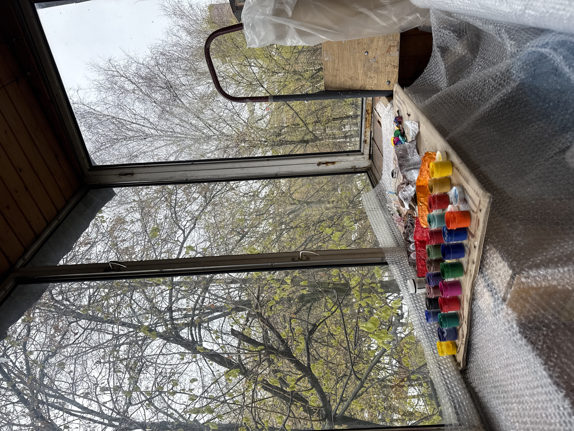 The image captures an indoor view looking out through a window, rotated 90 degrees clockwise. The perspective is from inside a room, possibly a sunroom, enclosed balcony, or a small shed-like structure, indicated by the wooden paneling visible in the upper left.
On the right side of the image, indoors, there's a surface covered extensively with bubble wrap, giving it a textured, reflective appearance. On this surface, positioned near the window, is a collection of small, colorful containers, likely paint pots or bottle caps, arranged in rows on a piece of light-colored cardboard or wood. These containers display a vibrant spectrum of colors including yellow, red, orange, blue, green, purple, and black. Other miscellaneous items, such as a white plastic bag and a curved metal bar (possibly part of a chair or rack), are also visible indoors.
The window itself is divided into multiple panes by metal or wooden frames, showing some signs of age or rust. Through the window, the outdoor scene is dominated by numerous deciduous trees. Most branches are bare, characteristic of late winter or early spring, but many show small clusters of fresh, light green leaves just beginning to unfurl, strongly suggesting the season is early spring. The sky beyond the trees is overcast and bright, indicating daytime with diffused light rather than direct sunlight. No people or animals are visible, and no specific activity is taking place. The scene appears calm and static. There is no visible text in the image.