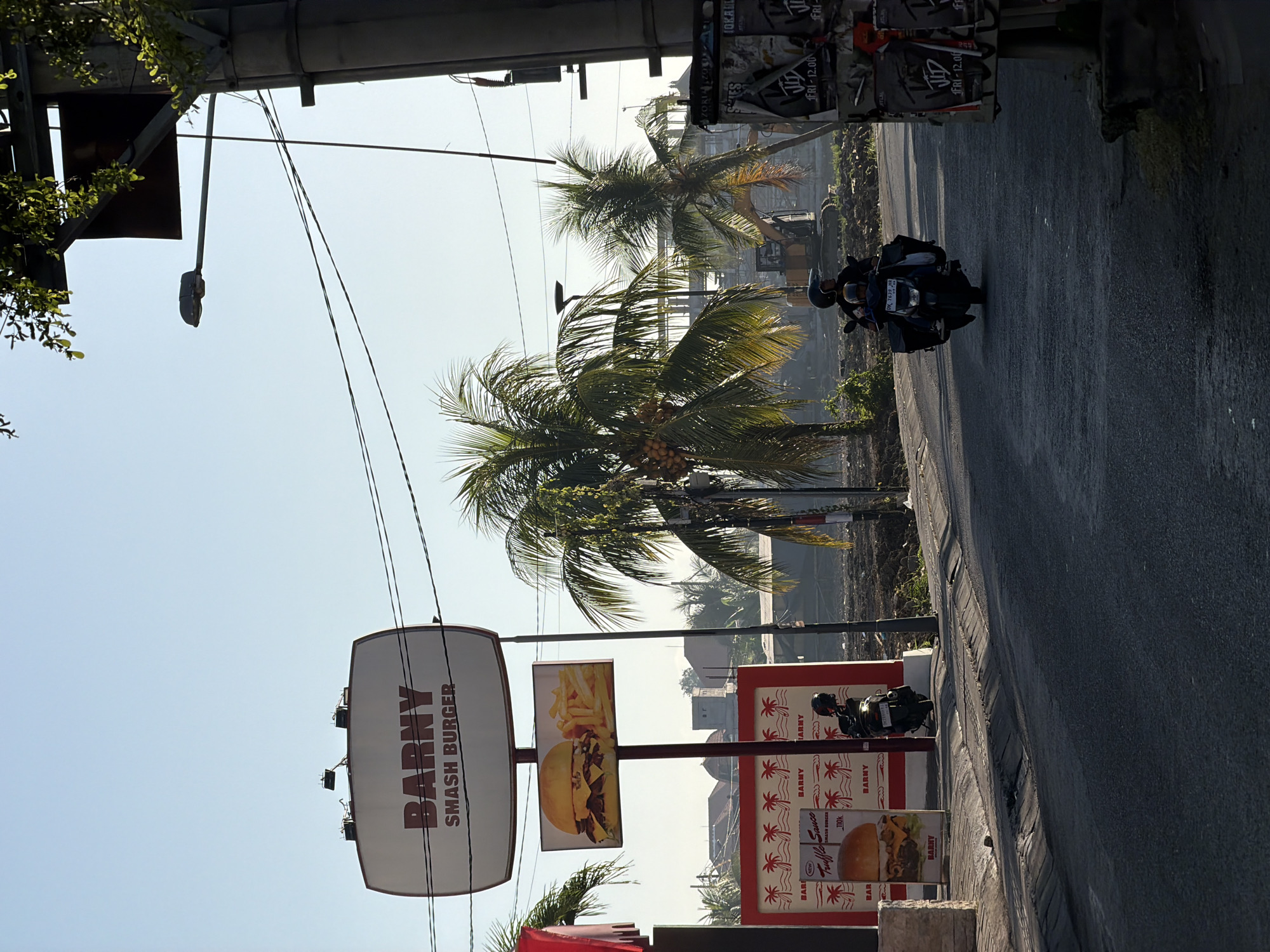 This is a street-level, medium-wide shot on a clear day in North Kuta, Indonesia. The foreground is dominated by a paved road with a gutter. On the left side of the road is a sidewalk with a red and white sign displaying "BARNY SMASH BURGER" with an image of a burger and fries. Next to it, a black scooter is parked. Further left, part of a building facade is visible. In the middle ground, a construction site is visible with an excavator. Several palm trees are scattered throughout the scene. On the right side of the road, a utility pole and various posters are mounted on a structure. Two people are riding a scooter in the middle of the road, traveling away from the viewer. The sky is clear and blue with some faint clouds. The lighting suggests it is morning or late afternoon.