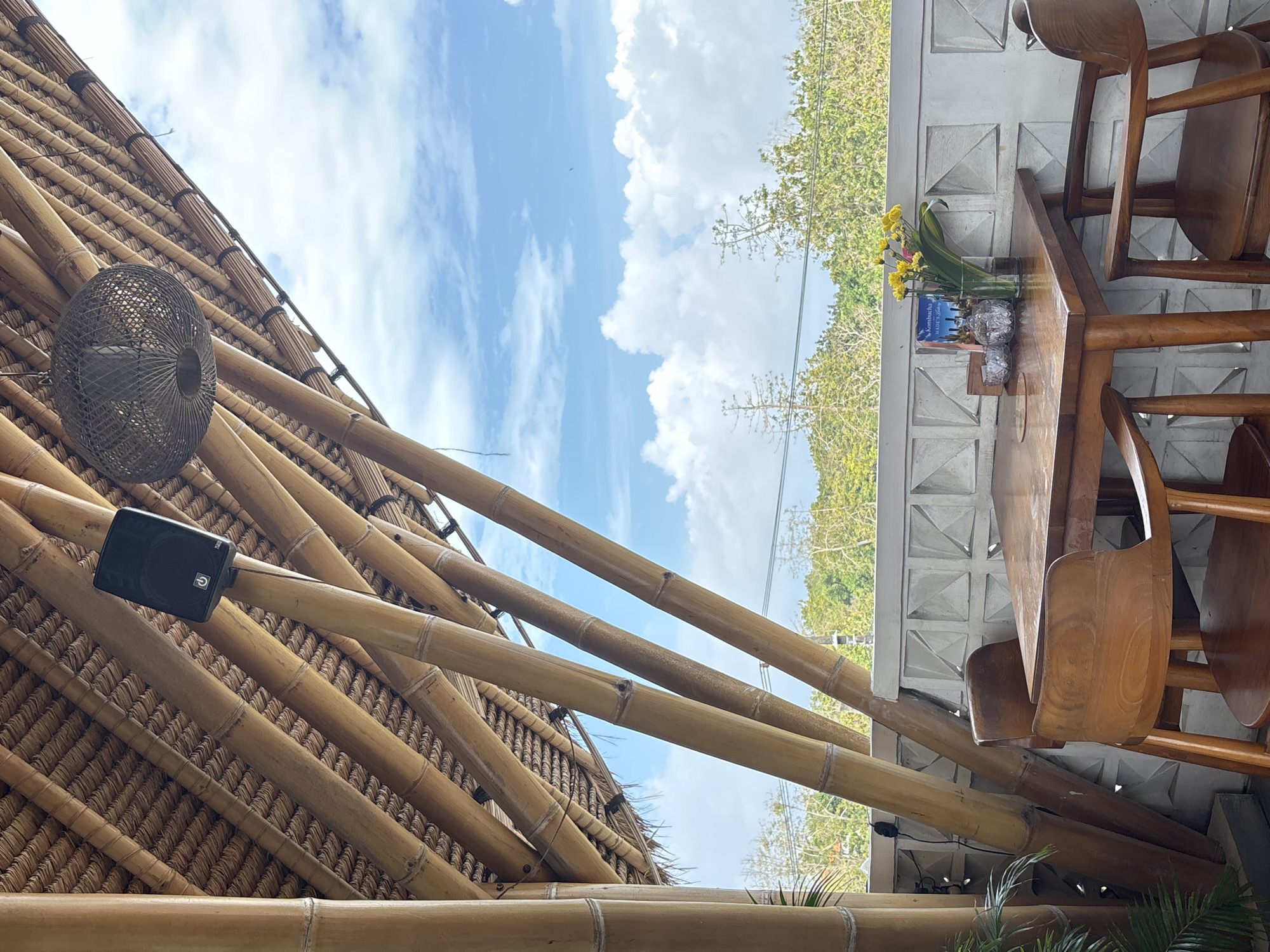 A table for two is set on a covered patio in Kuta Selatan, Indonesia. The patio features a bamboo ceiling and supports, with a woven light fixture and a speaker mounted overhead. The table is made of wood and has two wooden chairs with curved backs. On the table, there is a vase of yellow flowers, a small glass bowl, a menu, and a sugar dispenser. Beyond the patio railing, lush green trees and a cloudy blue sky are visible.