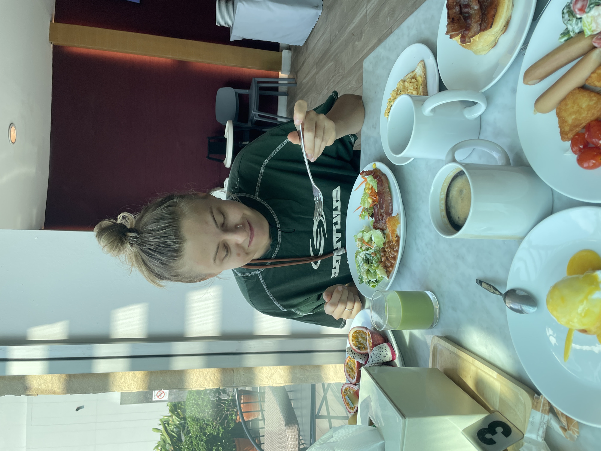 This image captures a bright daytime scene in a modern dining area, likely a hotel or restaurant in Bangkok, Thailand. A young woman with blonde hair tied in a bun is seated at a light-colored marble table, enjoying what appears to be a breakfast or brunch. She wears a dark green long-sleeved shirt with white lettering and a brown strap around her neck. She holds a fork, looking down with a content expression, suggesting an enjoyable meal.
The table is laden with various breakfast dishes. In front of the woman is a plate with a salad, bacon, and baked beans. To her right, a glass of green juice (possibly matcha or a vegetable blend) and a plate of vibrant, sliced tropical fruits, including passion fruit and dragon fruit, are visible. Other white plates hold an array of items: sausages, hash browns, scrambled eggs, cherry tomatoes, and a pastry topped with bacon. A white mug containing coffee and another empty white mug are also on the table. A white tissue box with a black number "3" on a clear block, likely a table number, is in the foreground.
Large windows behind the woman reveal an outdoor area with lush greenery and bright sunlight, indicating clear weather. A faint "no smoking" sign can be seen outside. The interior features a dark red wall accented by a wooden beam and subtle lighting. Dark grey chairs and a table covered with a white cloth are visible in the background, completing the ambiance of a pleasant dining experience.