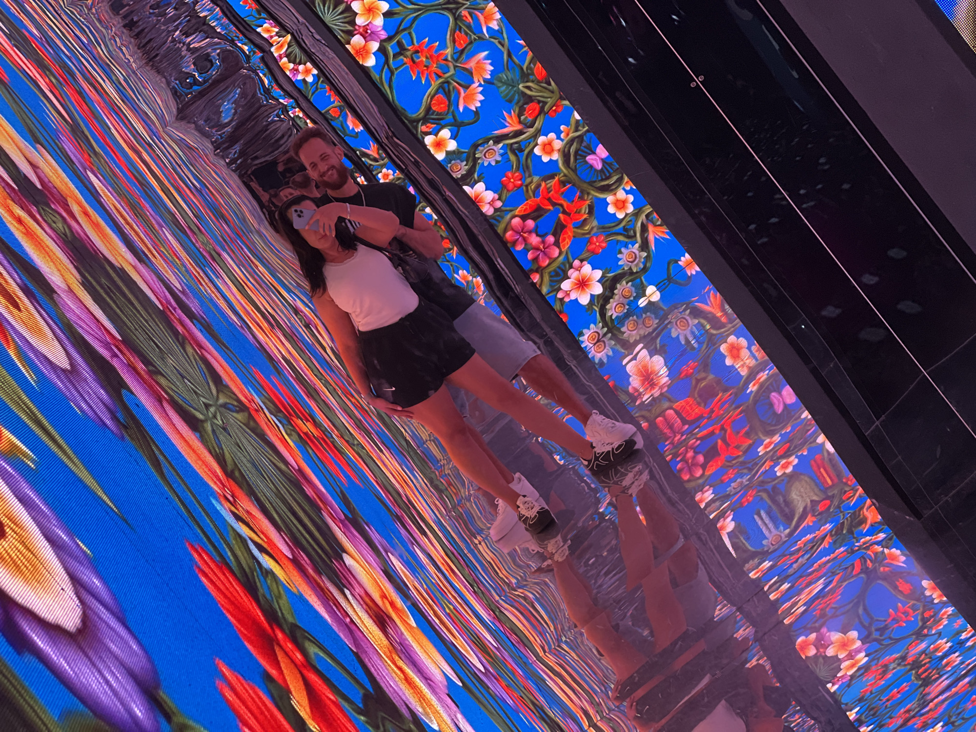 In Miami, Florida, a couple is taking a mirror selfie in a digital art installation. The woman, on the left, is wearing a white tank top and a black skirt. The man, on the right, is wearing a black t-shirt. They are standing on a reflective surface, which shows their reflections as well as the digital art installation that surrounds them. The installation features vibrant floral patterns and abstract designs in shades of blue, pink, red, and orange, creating a kaleidoscopic effect. The scene is brightly lit, and the overall atmosphere appears to be joyful and playful.