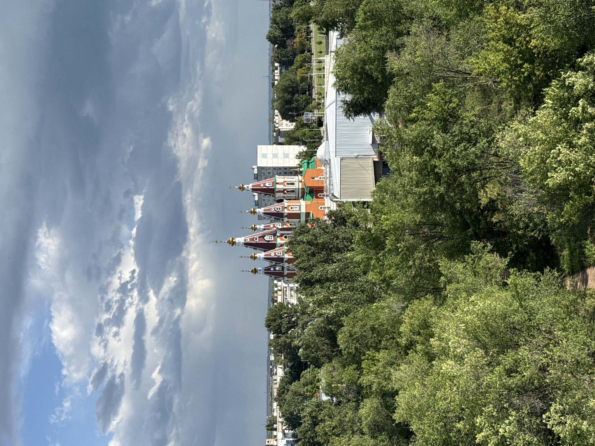 This is a high-angle, medium shot of a cityscape dominated by lush green trees. In the distance, a vibrant orange and red church with golden domes stands out against a backdrop of white and gray buildings under a dramatic, cloudy sky. The church has a traditional architectural style with multiple towers topped with crosses. The foreground is filled with dense foliage, suggesting a park or wooded area within the city. A narrow dirt path is visible winding through the trees on the right side of the frame. The sky is a mix of bright blue and dark, overcast clouds, hinting at dynamic weather conditions. The city of Otradny, Russia, is visible in the background, with the church being a prominent landmark within it.