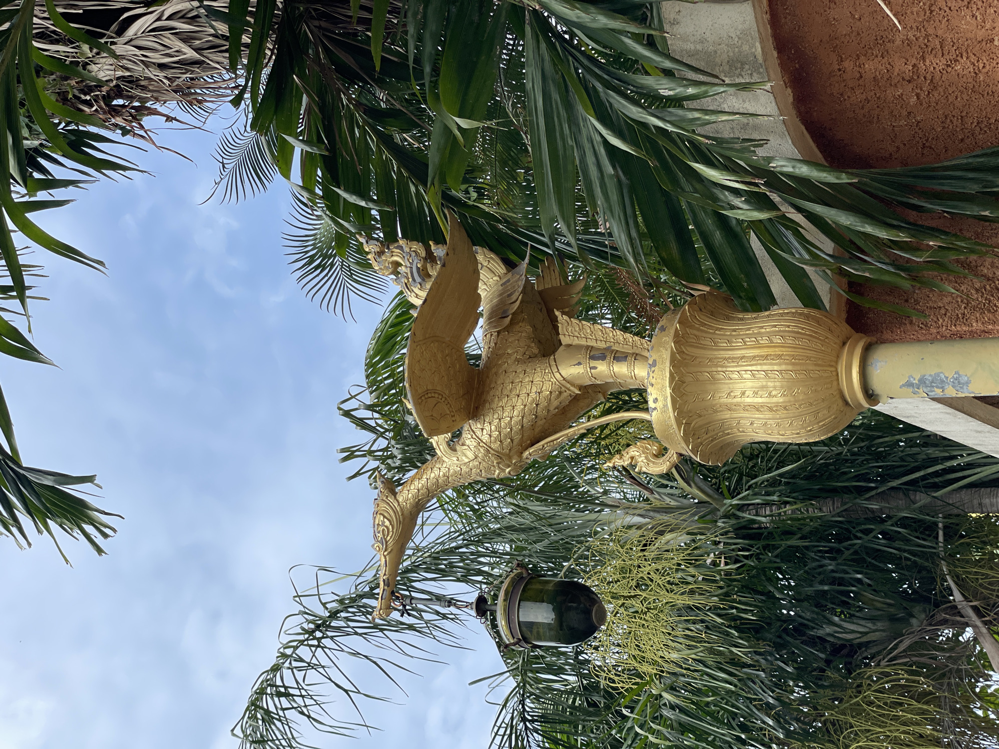 An ornate golden bird statue with outstretched wings and a long neck stands prominently in front of lush green palm trees under a cloudy blue sky. A dark green glass lantern hangs from the statue's beak by a metal chain. The statue is mounted on a golden decorative pedestal that is attached to a weathered, light-colored pole. The background consists of dense palm fronds and a brown textured wall. This scene is located in Pattaya City, Thailand, likely within a temple or a garden area, given the artistic style of the statue and the tropical vegetation. There are no people or animals visible in the image.