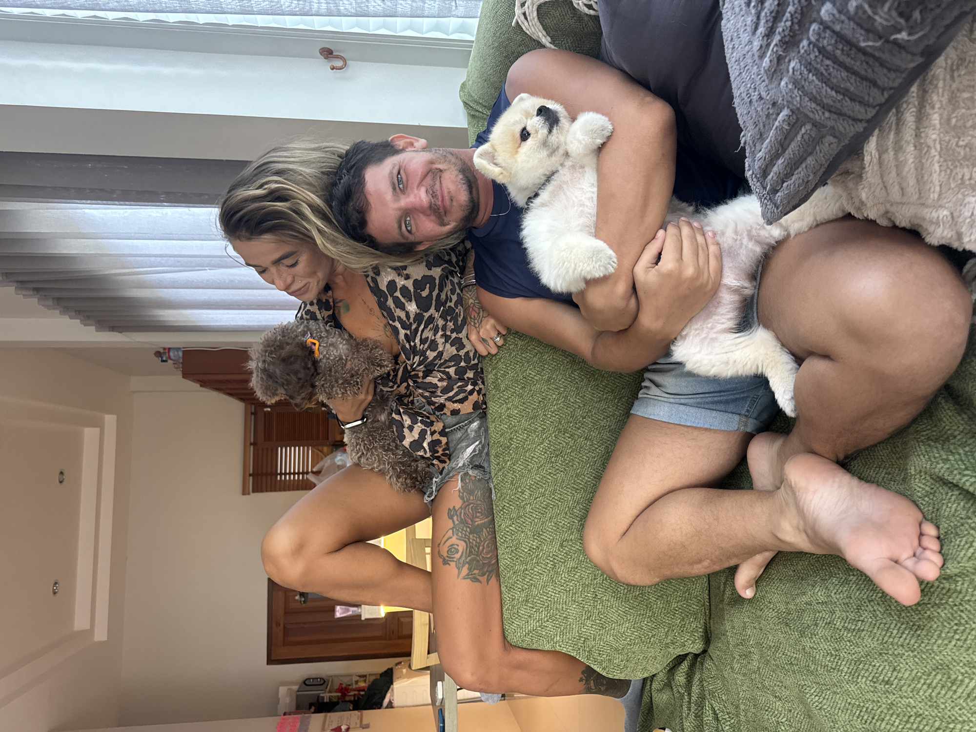 In the city of Thep Krasatti, Thailand, a man and a woman are relaxing on a green sofa. The woman is reclined on the back of the sofa, with her legs bent and crossed. She is holding a small, dark-colored poodle in her arms. She is wearing a leopard-print top and denim shorts. The man is seated on the sofa, holding a small, white dog, likely a Pomeranian, in his arms. He is wearing a dark blue sleeveless shirt and denim shorts. Both individuals appear relaxed and are looking towards each other. The room is well-lit, with natural light coming from a large window with sheer curtains on the right side of the frame. Behind the sofa, there is a wall with what appears to be a framed picture or artwork. The environment suggests a comfortable and casual home setting.