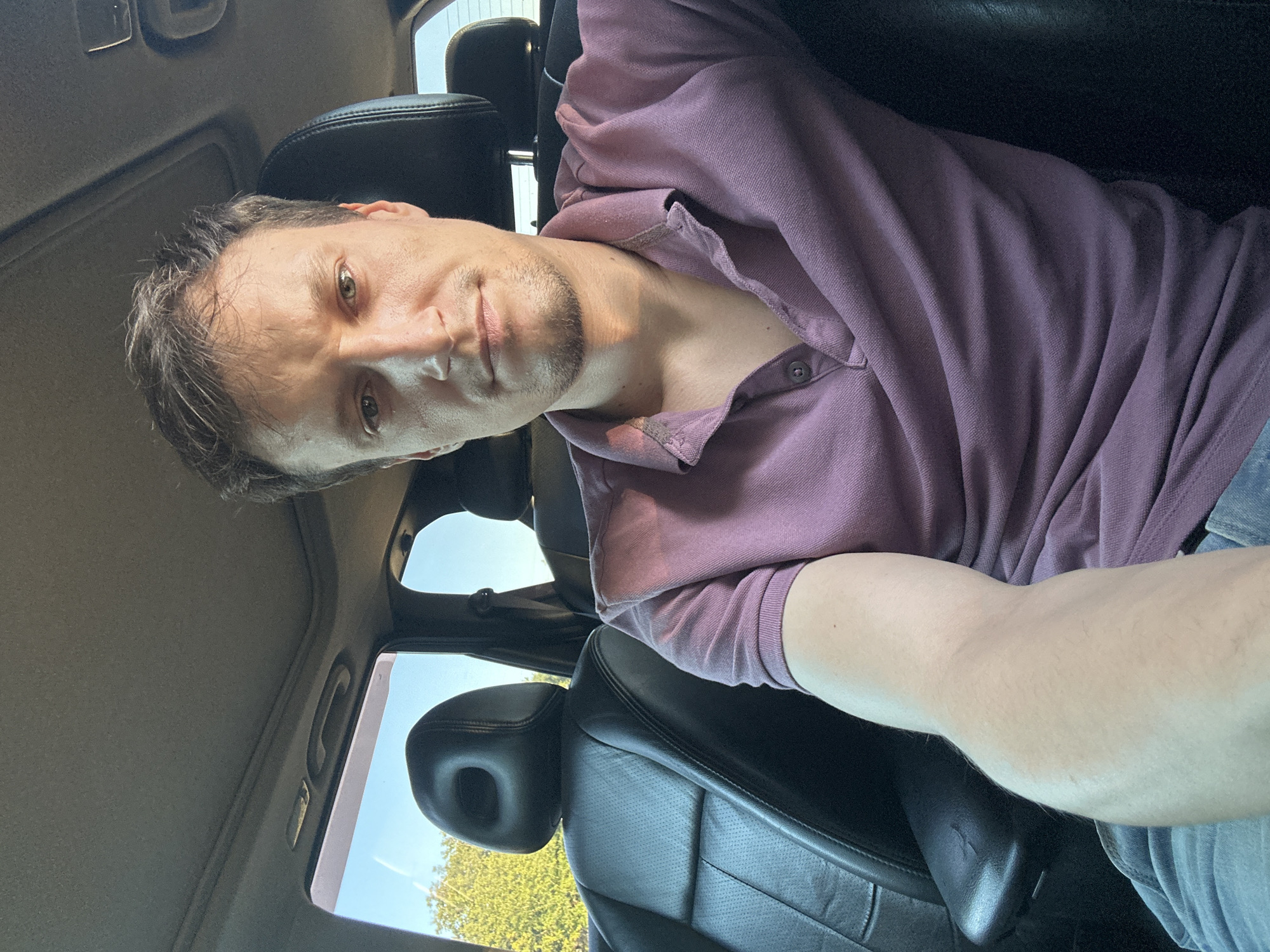 The image is a selfie taken from the driver's seat of a car, likely in the city of Makiv, Ukraine, given the context. The subject, a man with light brown hair and a light beard, is looking directly at the camera. He is wearing a purple polo shirt. The interior of the car is visible, including black leather seats, the headrest of the seat next to him, and the car's headliner with a sunroof. Sunlight streams into the car from the windows, illuminating the scene. Outside the car, green trees and a blue sky are visible.