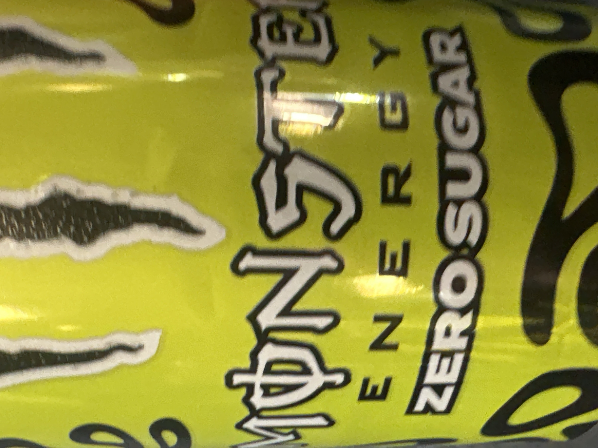 The image shows part of a yellow can of Monster Energy Zero Sugar drink. The words "Monster Energy Zero Sugar" are visible. The can has a black claw mark design on the surface.