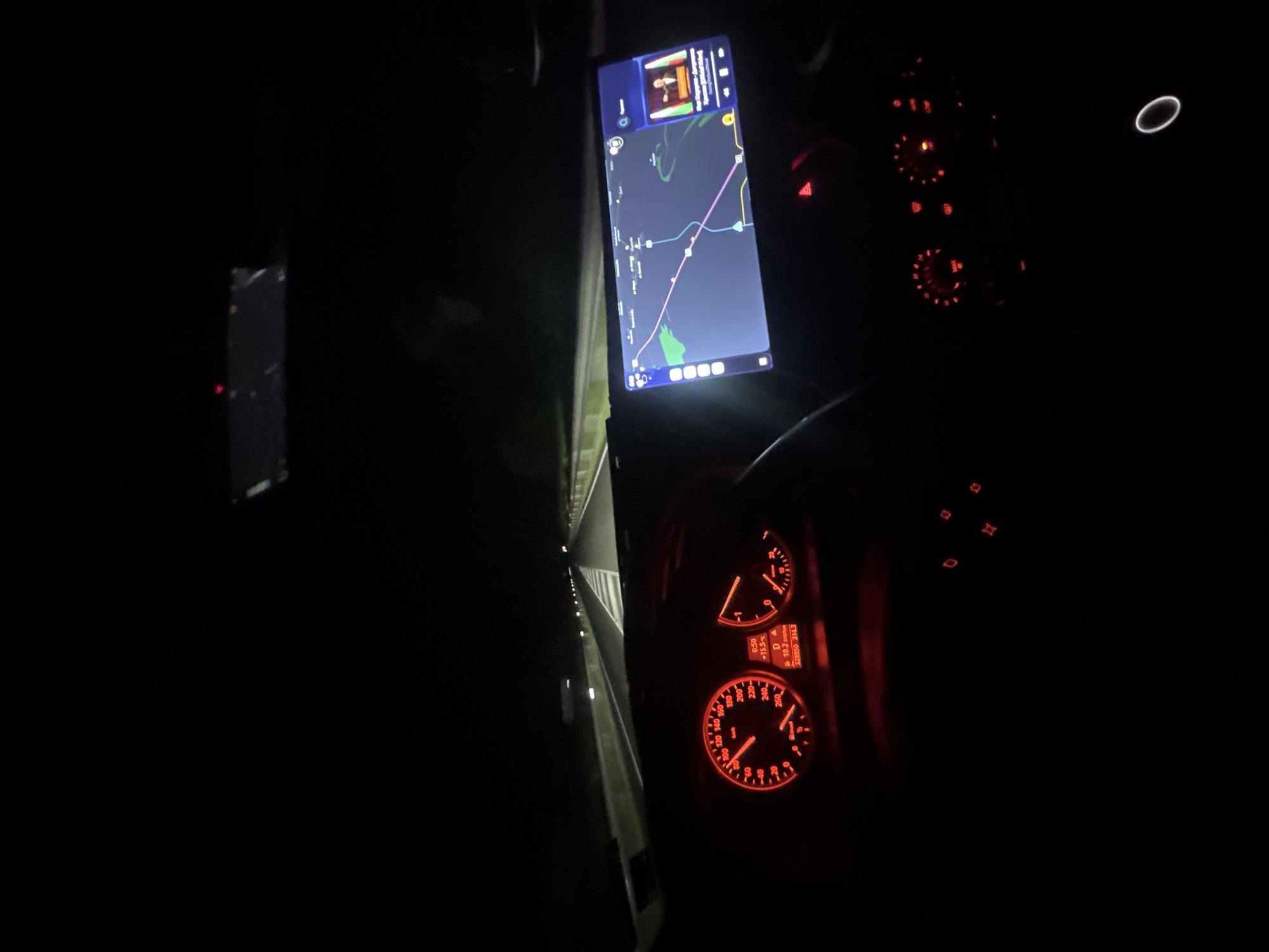 The image shows the interior of a car at night, likely while driving. The speedometer and other dashboard gauges are illuminated in red. A navigation app is displayed on a screen mounted near the dashboard, showing a map with a route marked in purple. The location is likely in Mogila, Bulgaria based on the map displayed on the navigation app. The image captures a dark environment with focus on the driver's view of the road and navigational aids.
