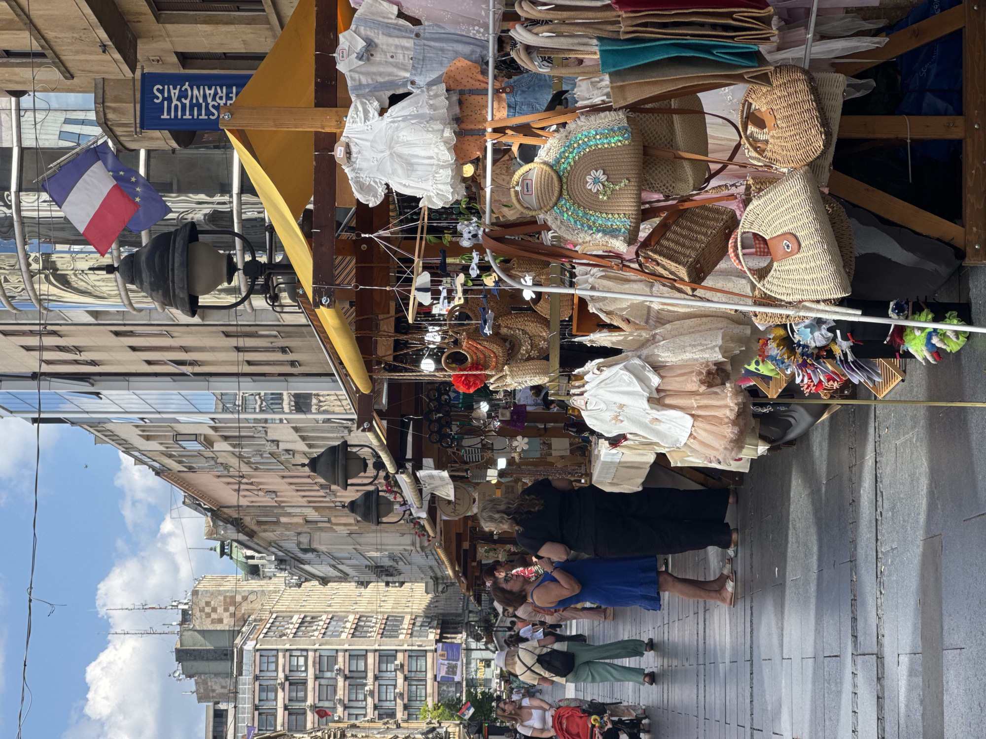 This is a vertical, eye-level, medium shot of a street market stall in Belgrade, Serbia. The stall displays a variety of items, including clothing, handbags, and accessories. Several people are browsing the merchandise, suggesting a shopping scene. The time of day appears to be afternoon, with the sun casting shadows on the cobblestone street. The sky is partly cloudy, indicating fair weather.
The stall is adorned with a yellow awning and features racks of clothes and shelves filled with woven bags. Among the notable objects are several straw handbags, some decorated with colorful beads and floral patterns, and a white lace dress hanging prominently. A French flag and the blue sign "INSTITUT FRANÇAIS" are visible, indicating the proximity of the French Institute.
In the foreground, several individuals are seen walking and examining the goods. One woman in a blue dress stands out, and another in a black top appears to be a vendor. The architecture of the surrounding buildings, characterized by their multi-story structures and windows, suggests an urban setting. The overall atmosphere seems relaxed and casual, typical of a daytime street market. The image captures a moment of everyday life and commerce in a European city.