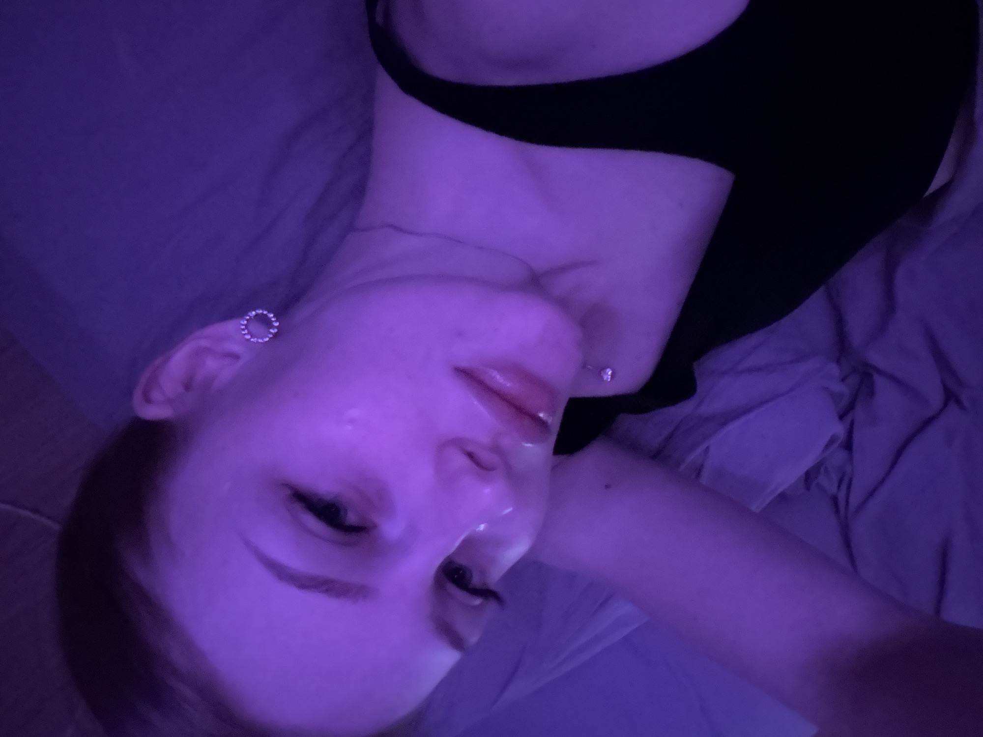 A person with light skin and dark hair is lying on a bed. The person is wearing a black top, and there is an earring in their left ear and a piercing on their neck. The lighting is blue and purple. The location is not identifiable as Perm, Russia, from the given information.