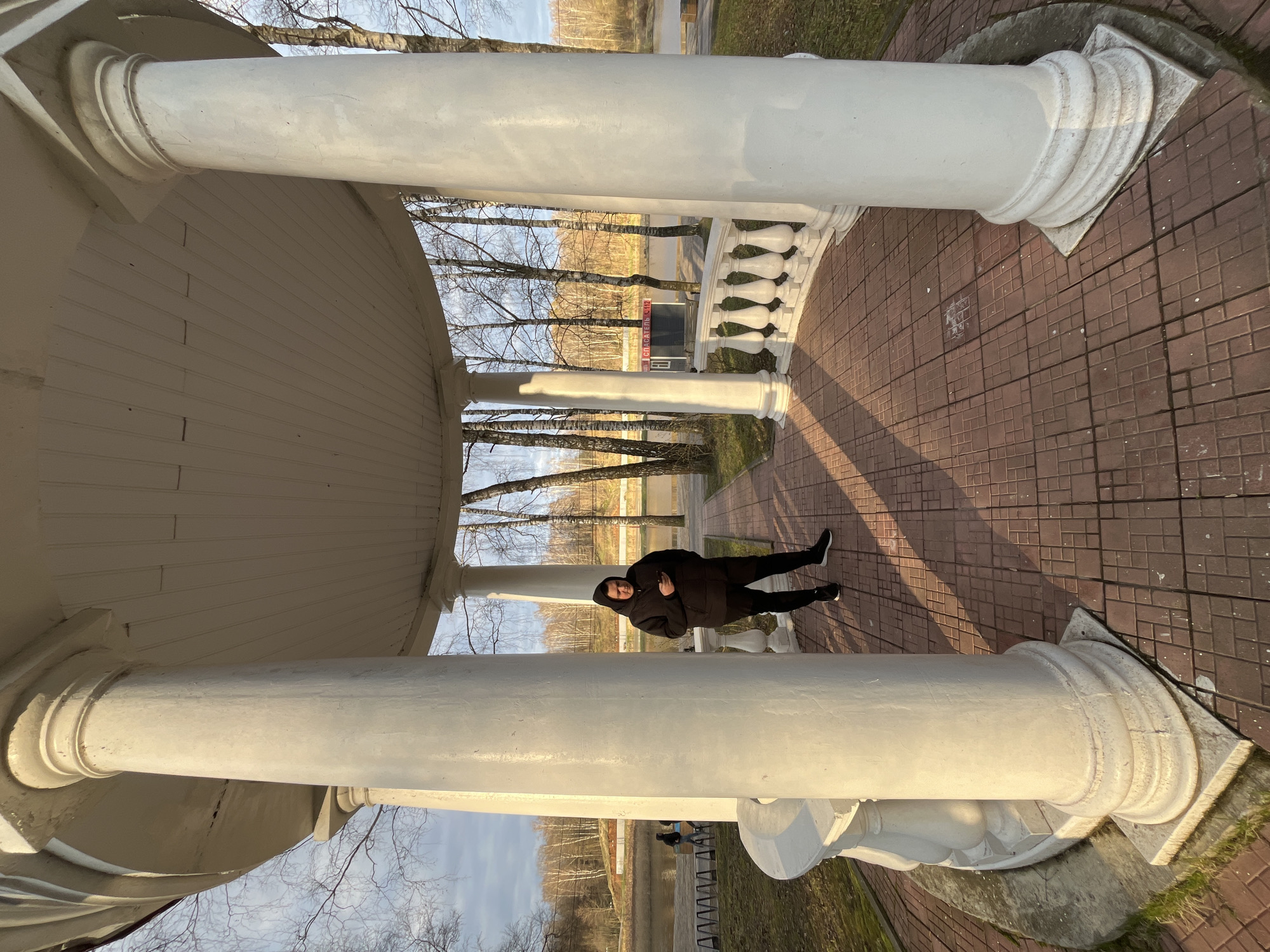 A medium, eye-level shot shows a person standing within a white, columned gazebo-like structure, likely in the city of Klin, Russia. The structure has a tiled floor of reddish-brown bricks arranged in a square pattern. The gazebo has at least four white, cylindrical columns with decorative bases and capitals. The person is wearing a dark-colored puffy jacket, dark pants, and black athletic shoes. The person stands with their arms crossed. Beyond the structure, there is a view of bare trees, grass, and a building with Cyrillic lettering. The lighting suggests it is daytime with sunshine.