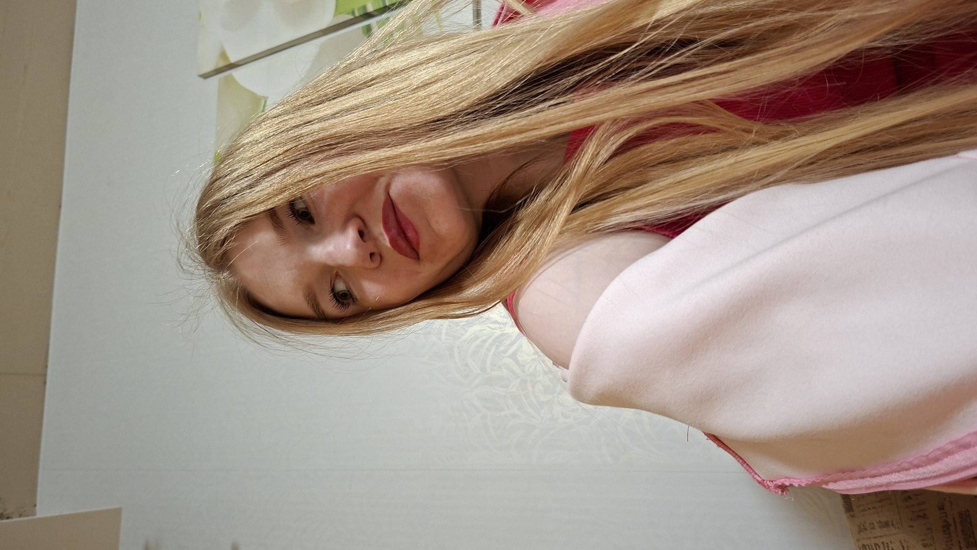 This is an upright portrait photograph of a young person, likely female, captured from a slightly low angle, giving a dynamic perspective. The subject's head is tilted and rotated, featuring long, straight, blonde hair that sweeps across the upper part of the frame. She has light-colored eyes, prominent eyelashes, and is wearing dark red or deep pink lipstick, with a subtle, slightly pouting or playful expression.
She is dressed in a pink garment, partially visible on her right side, and a lighter, possibly off-white or pale pink, soft fabric—appearing like a blanket or robe—is draped over her right shoulder and arm.
The background suggests an indoor setting. A light-colored wall with a subtle, textured floral or damask wallpaper pattern is visible. Above the subject's head, there's a hint of a framed picture or mirror reflecting green and white floral designs. In the bottom right corner, a small, blurry piece of paper with illegible printed text, possibly from a newspaper or product packaging, is visible on a surface.
The lighting appears bright and natural, indicating a daytime shot. The overall impression is casual and personal. The image's location context is Большой Букор, Russia.