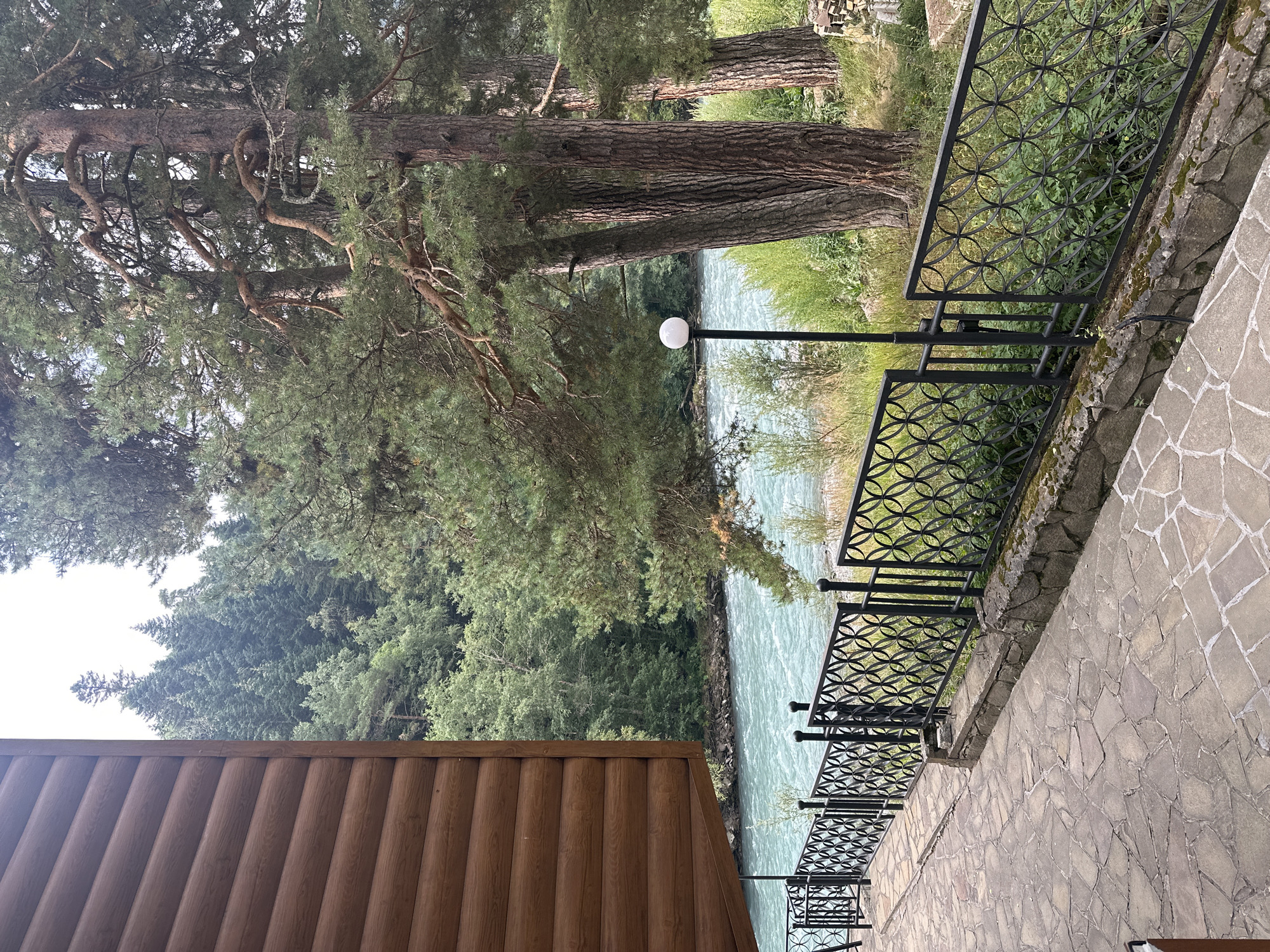 A picturesque scene unfolds with a rustic wooden building on the left, its horizontal planks forming a textured wall. A paved walkway, laid with irregular stone slabs, leads towards a black metal railing adorned with circular patterns. Beyond the railing, a vibrant turquoise river rushes by, its surface rippling with dynamic movement. Lush green trees, predominantly pines with their characteristic needle-like foliage and gnarled branches, dominate the background. Their tall trunks stand sentinel, some appearing weathered and mature. A solitary, spherical white lamp on a dark pole stands near the riverbank, providing a touch of artificial light amidst the natural splendor. The environment suggests a serene, natural setting, likely a resort or lodge area in a mountainous region. The scene is devoid of people. The overall impression is one of tranquility and the harmonious coexistence of human construction with the raw beauty of nature, evoking a sense of peace and escape. The location is likely the resort area in Arkhyz, Russia, known for its stunning natural landscapes and outdoor recreational opportunities.