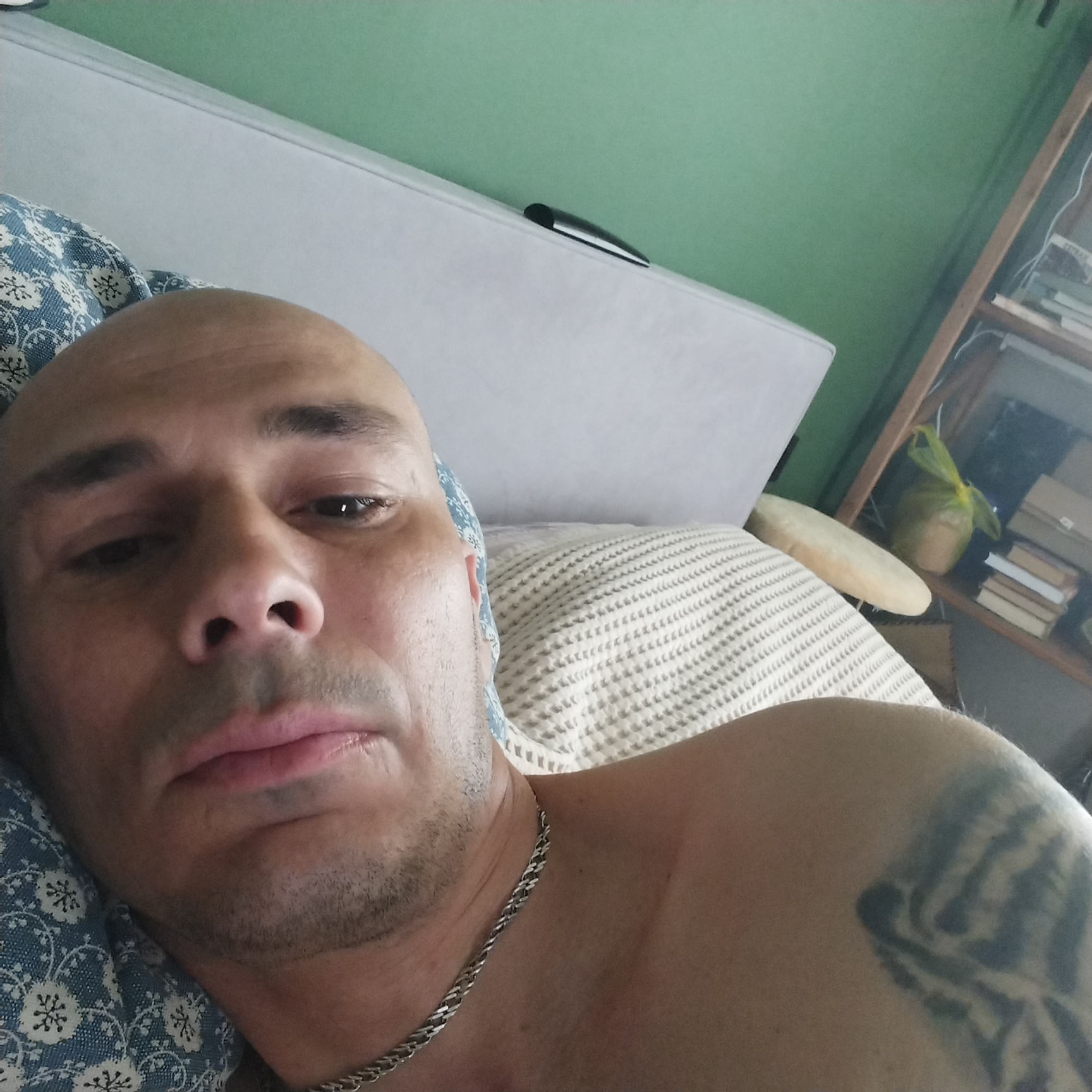 A bald man with light skin and a short beard is lying on a bed, looking towards the camera. He has a tattoo on his upper right arm and is wearing a silver chain necklace. The room has a green wall, a white bed with a patterned blue and white pillow, and a wooden bookshelf filled with books and other items. A yellow bag is visible on the shelf. The scene appears to be indoors, in a residential setting. The provided metadata indicates the image was taken in Vladivostok, Russia.