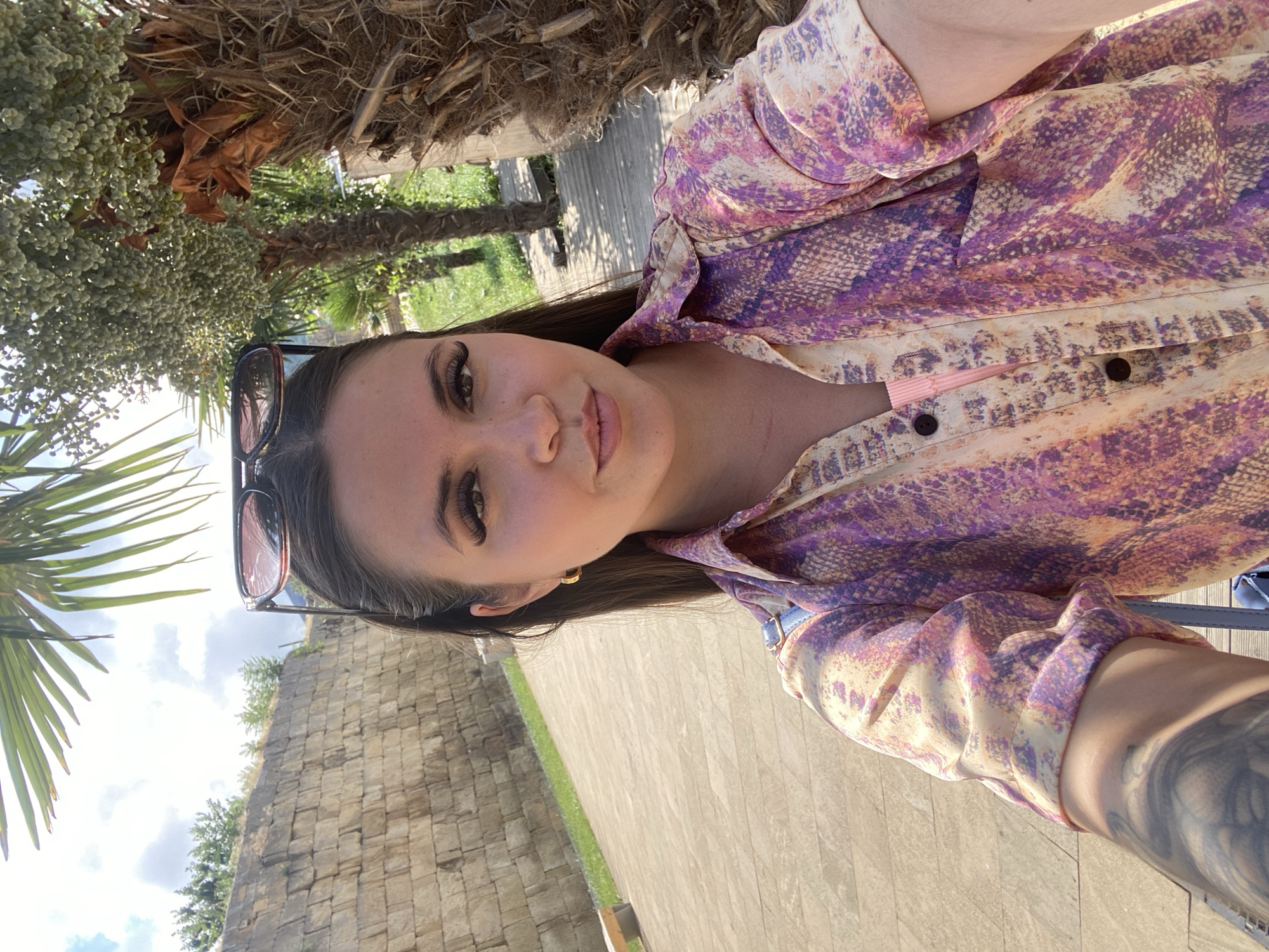 A young woman with dark hair pulled back and sunglasses perched on her head takes a selfie outdoors. She is wearing a colorful patterned shirt over a pink garment. Her left arm, tattooed, is visible in the foreground. The background features a stone wall, palm trees, and a pathway. The location is likely Derbent, Russia, given the context provided.