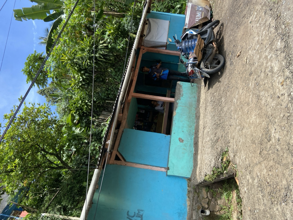 The image shows a small, blue building with a wooden awning located in Cipatat, Indonesia. The building is set into a hillside. There is lush vegetation above and behind the building. There are two people, both wearing blue shirts, standing near the front of the building. One person is holding a black and white bag and the other is holding a blue bag. A black motorcycle is parked on the dirt road in front of the building. The motorcycle has a blue water bottle in its side basket and a brown cardboard box on its rear rack. There is a sign with the text "UMI" in white on a red background behind the motorcycle. There is a small drain in the foreground of the image, leading to a dry stone-lined ditch.