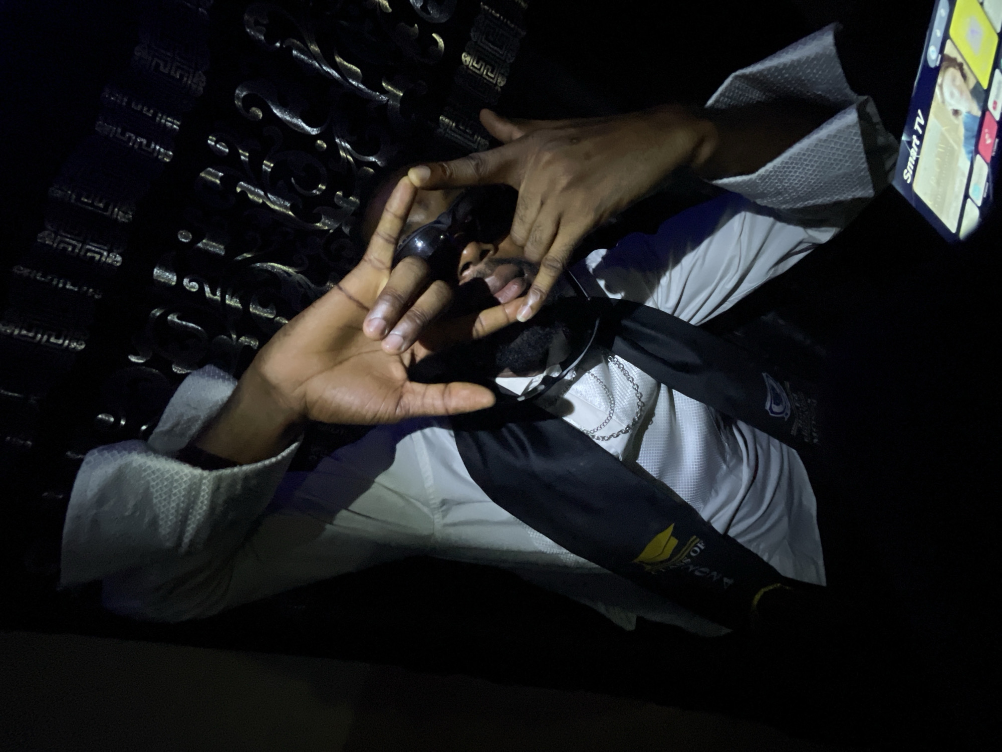 A person sits in dim lighting, possibly indoors. They are wearing a light-colored, patterned shirt under a black graduation gown with yellow accents. Their hands are positioned in front of their face, forming a triangular shape. A smartphone displaying a screen with app icons is visible in the upper right corner. The background includes a patterned surface, possibly upholstery or decorative paneling. The exact location in Lokoja, Nigeria, is not evident from the image.