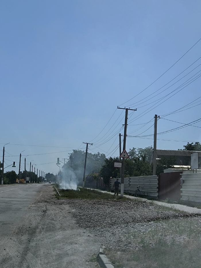 The image displays a daytime street scene in Illintsi, Ukraine, under a clear blue sky. A two-lane asphalt road, showing signs of wear and featuring loose gravel patches on its surface and shoulder, extends from the foreground towards the background on the left. A prominent plume of white smoke rises from ground level on the right side of the road, within a grassy median strip or shoulder area that contains loose gravel.
Multiple wooden utility poles with numerous overhead power lines and cables are visible, traversing the scene from left to right. Streetlights are present along the left side of the roadway. On the right, a dark-clothed individual, appearing male, walks away from the camera, partially obscured by a utility pole. A bicycle is propped against a corrugated metal fence section located behind the walking individual. A triangular road warning sign depicting two vehicles and a rectangular white sign with the Cyrillic text "БЕРЕЖИСЬ АВТОМОБИЛЯ" (Beware of Cars) are affixed to a utility pole on the right. Trees and a building with a dark roof are partially visible beyond the fences on the right side of the frame.