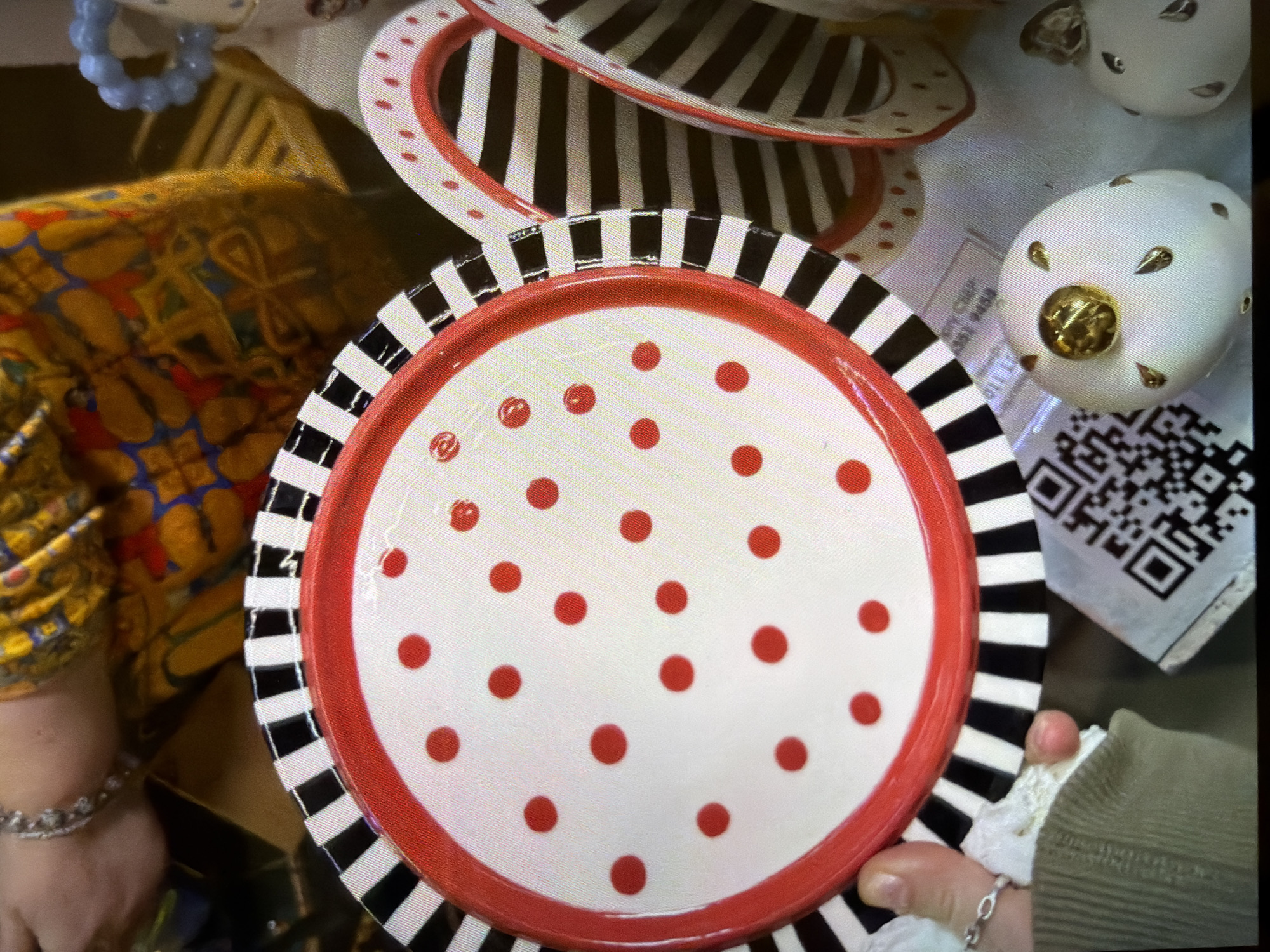 The image shows a stack of decorative plates. The plate in the foreground is white with red polka dots, a red rim, and a black and white striped border. Another plate with black and white stripes is partially visible behind it. To the right of the plate is a white decorative egg with gold accents, next to a small card with a QR code. A person wearing a mustard-yellow patterned shirt and jewelry is partially visible on the left. Another person's hand is holding the plate on the right. The location is not discernible in the image.