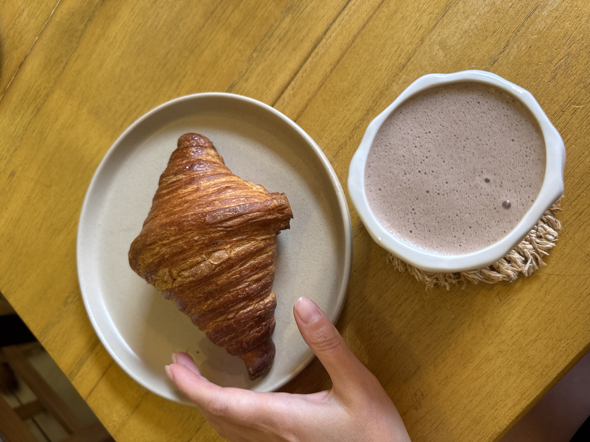 A hand is reaching for a croissant on a plate. The croissant is golden brown and flaky. A cup of hot chocolate is next to the plate. The scene is set on a wooden table. The image is taken from a high angle, looking down at the table. There is no indication of the location in Batumi, Georgia.