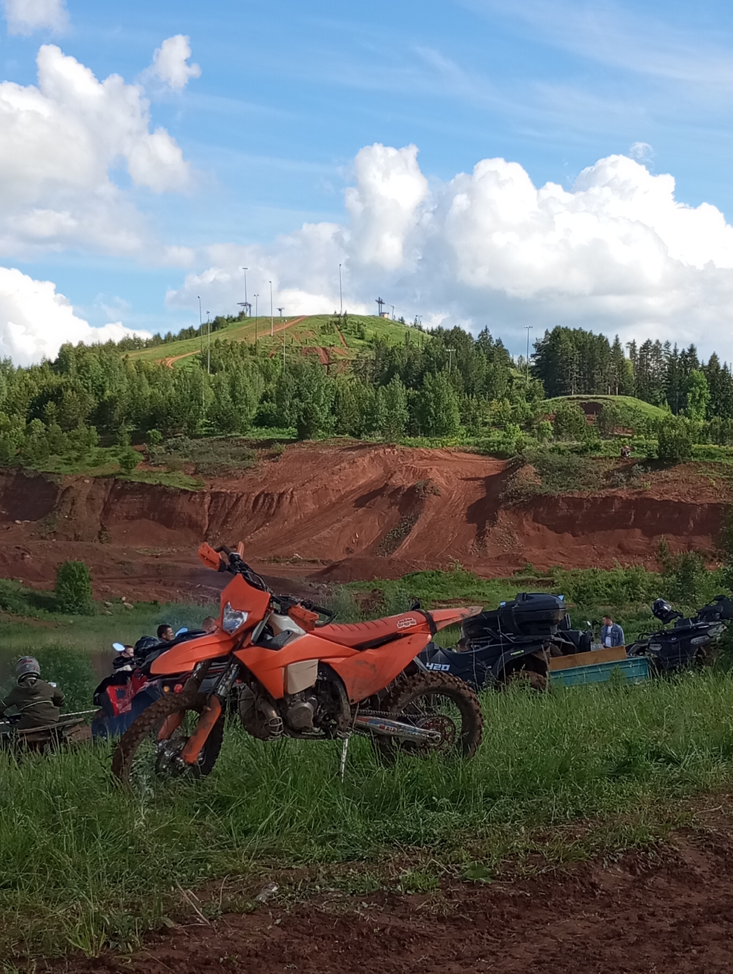 The image shows a dirt bike parked in a grassy area in front of a red dirt hill with tire tracks. Behind the dirt bike are several all-terrain vehicles (ATVs) and a group of people. In the background is a hill covered in trees, with ski lifts visible on the summit. The sky is blue with white clouds. This scene is located near the Chekeril ski resort in Izhevsk, Russia, based on the terrain and ski lift structures visible.